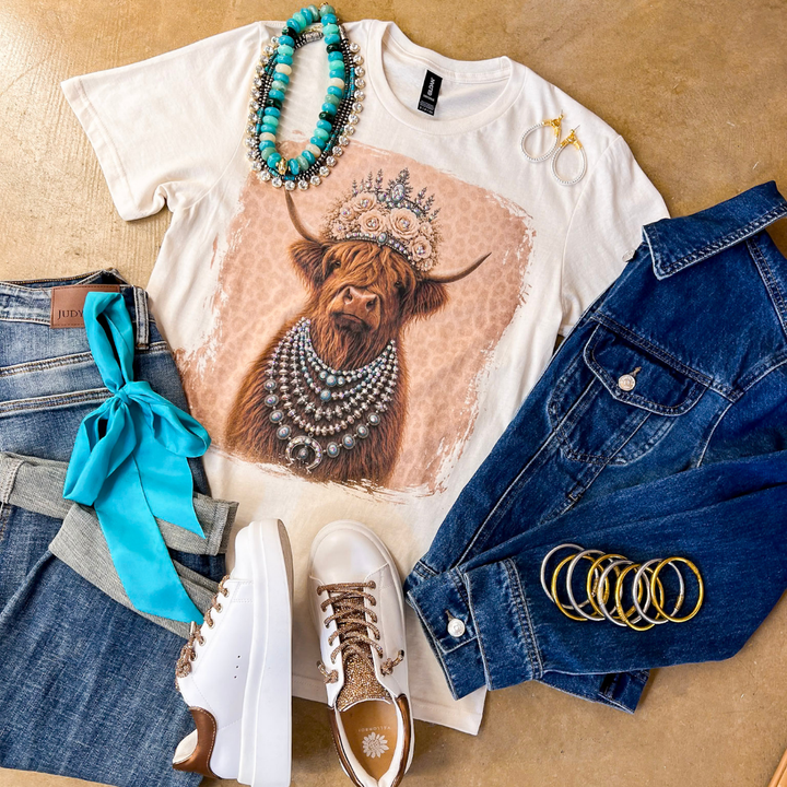 Heidi the Highland Queen crystal graphic tee in cream with a highland cow wearing a jeweled floral crown and layered necklaces on a pink leopard background, styled with denim and turquoise jewelry