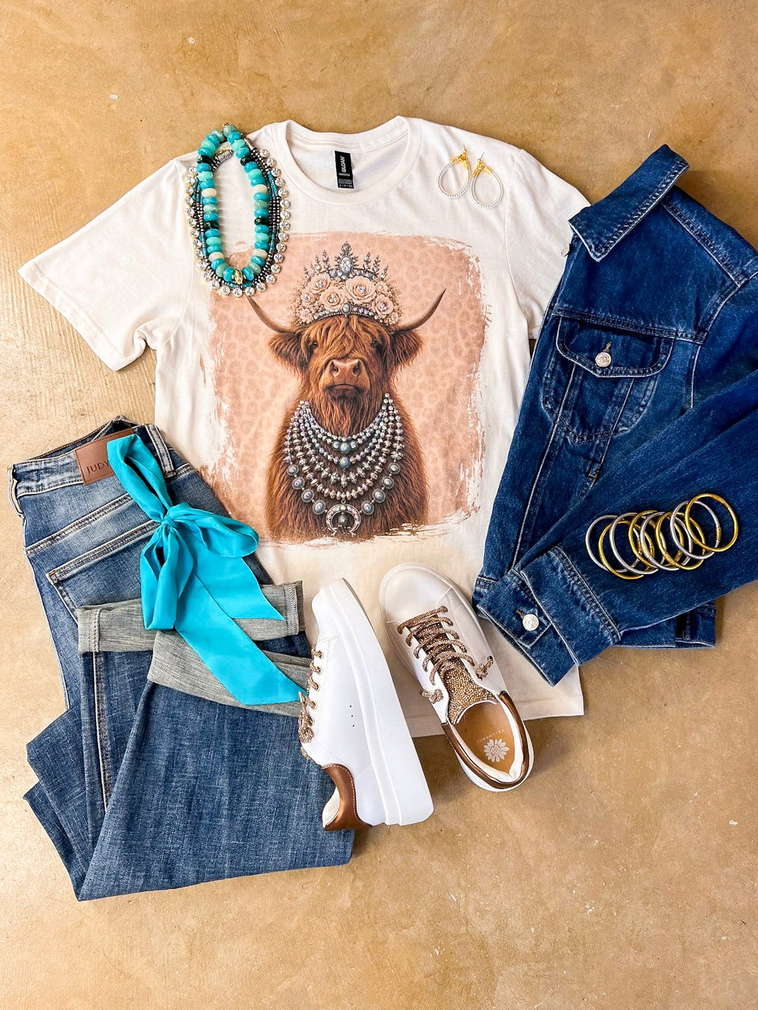 Heidi the Highland Queen crystal graphic tee in cream with a highland cow wearing a jeweled floral crown and layered necklaces on a pink leopard background, styled with denim and turquoise jewelry