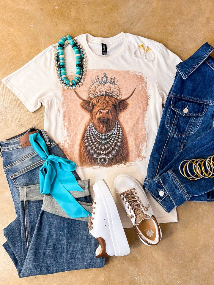 Heidi the Highland Queen crystal graphic tee in cream with a highland cow wearing a jeweled floral crown and layered necklaces on a pink leopard background, styled with denim and turquoise jewelry