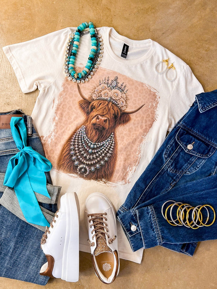 Heidi the Highland Queen crystal graphic tee in cream with a highland cow wearing a jeweled floral crown and layered necklaces on a pink leopard background, styled with denim and turquoise jewelry