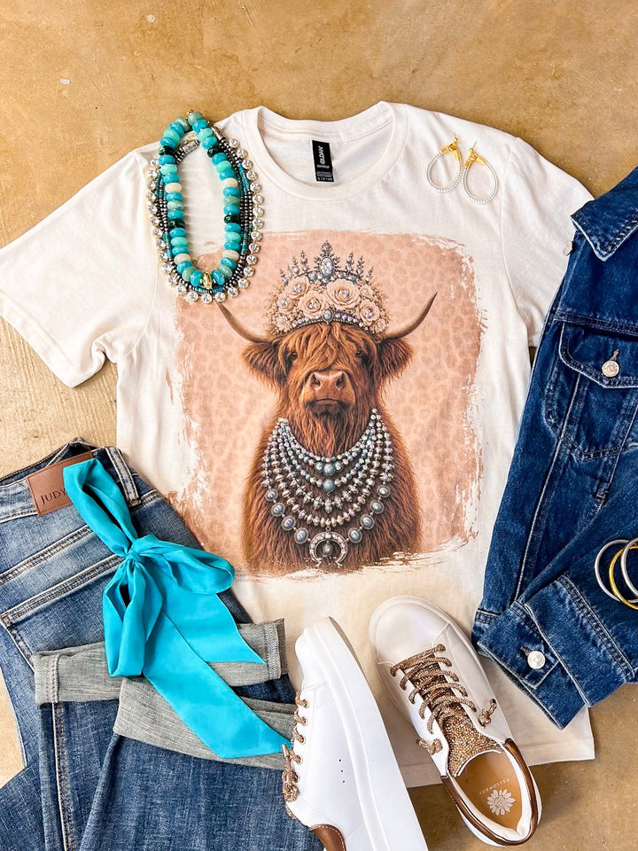 Heidi the Highland Queen crystal graphic tee in cream with a highland cow wearing a jeweled floral crown and layered necklaces on a pink leopard background, styled with denim and turquoise jewelry