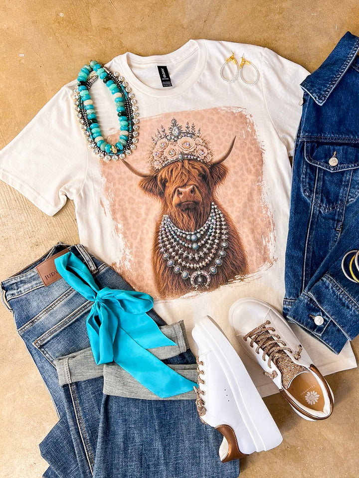 Heidi the Highland Queen crystal graphic tee in cream with a highland cow wearing a jeweled floral crown and layered necklaces on a pink leopard background, styled with denim and turquoise jewelry