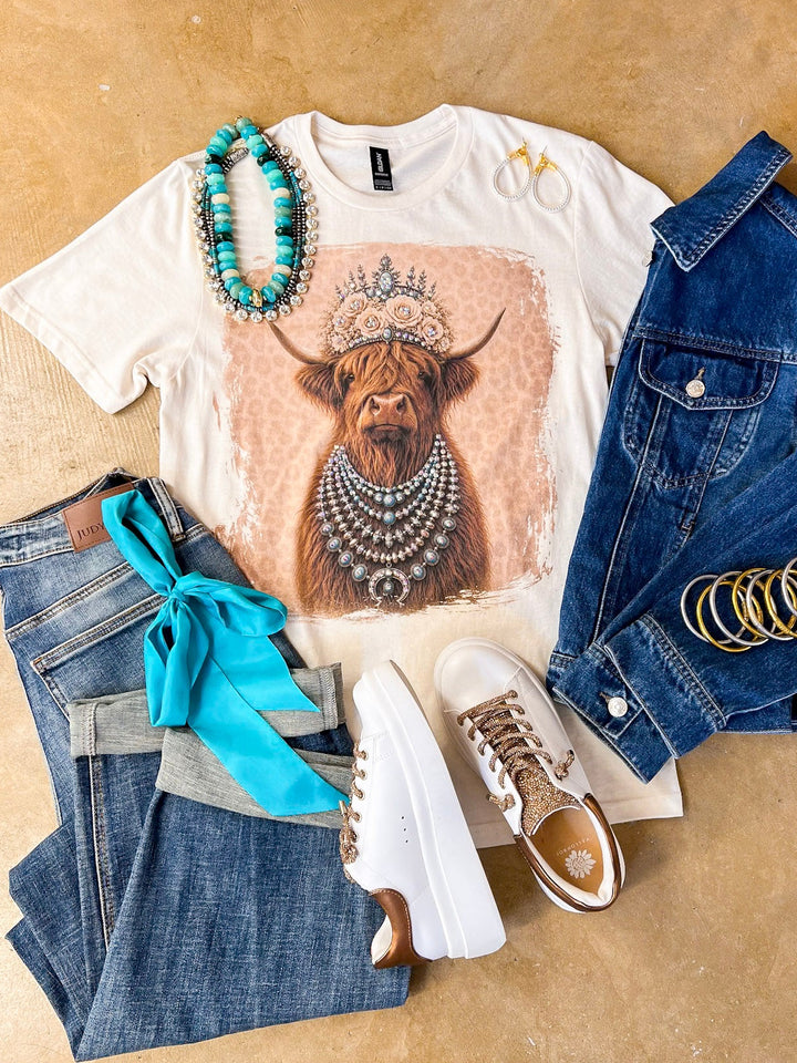 Heidi the Highland Queen crystal graphic tee in cream with a highland cow wearing a jeweled floral crown and layered necklaces on a pink leopard background, styled with denim and turquoise jewelry
