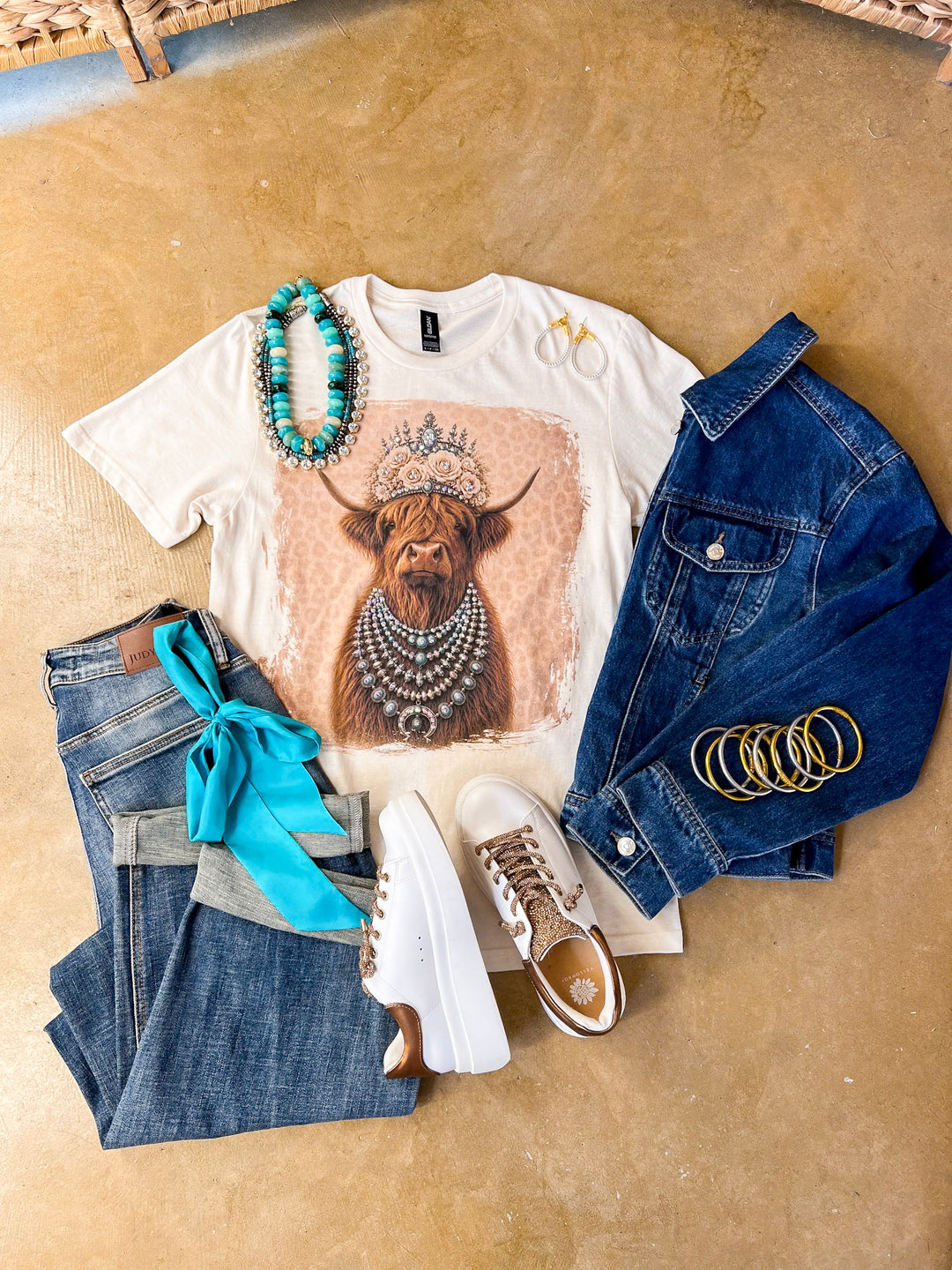Heidi the Highland Queen crystal graphic tee in cream with a highland cow wearing a jeweled floral crown and layered necklaces on a pink leopard background, styled with denim and turquoise jewelry