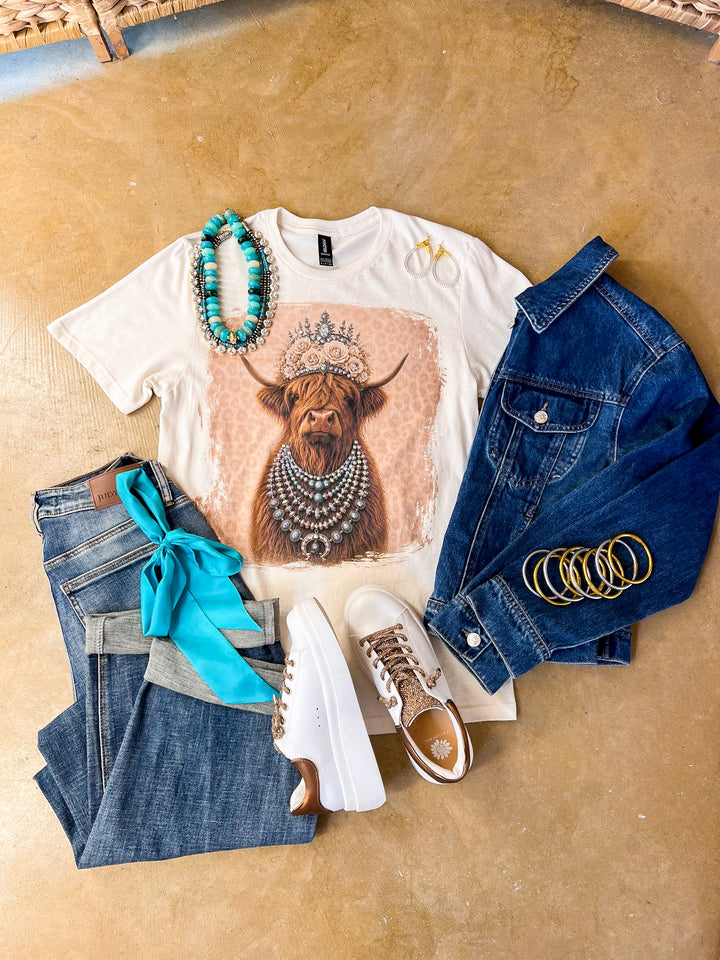 Heidi the Highland Queen crystal graphic tee in cream with a highland cow wearing a jeweled floral crown and layered necklaces on a pink leopard background, styled with denim and turquoise jewelry
