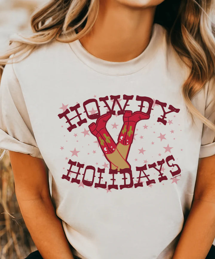 Cream “Howdy Holidays” graphic tee featuring red cowboy boots and pink stars — western Christmas shirt on model.