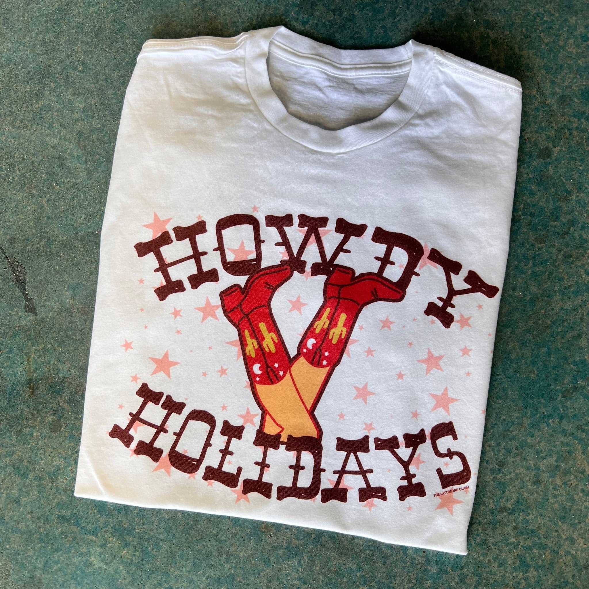 This white tee includes a crew neckline, short sleeves, and a hand drawn Christmas design of legs with red cowboy boots on. The cowboy boots have cactus and moon details on them, with pink stars all around the design, and the words "HOWDY HOLIDAYS" are above and below the design. The Western font is a fun touch to this graphic tee. This is shown as a folded flatlay on a blue background.