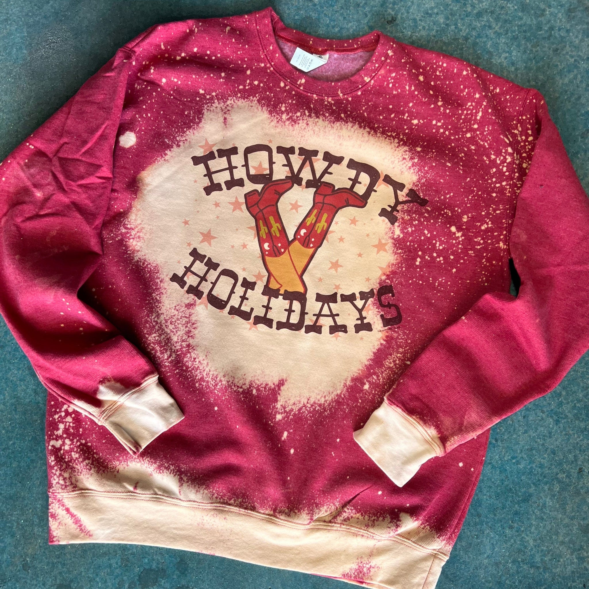 This red sweatshirt is bleached and includes a crew neckline, long sleeves, and a hand drawn Christmas design of legs with red cowboy boots on. The cowboy boots have cactus and moon details on them, and the words "HOWDY HOLIDAYS" are above and below the design. The Western font is a fun touch to this sweatshirt. The bleached look can be found all over the sweatshirt, bottoms of sleeves, bottom of sweatshirt, and behind the design. This is shown as a flatlay on a blue background. 