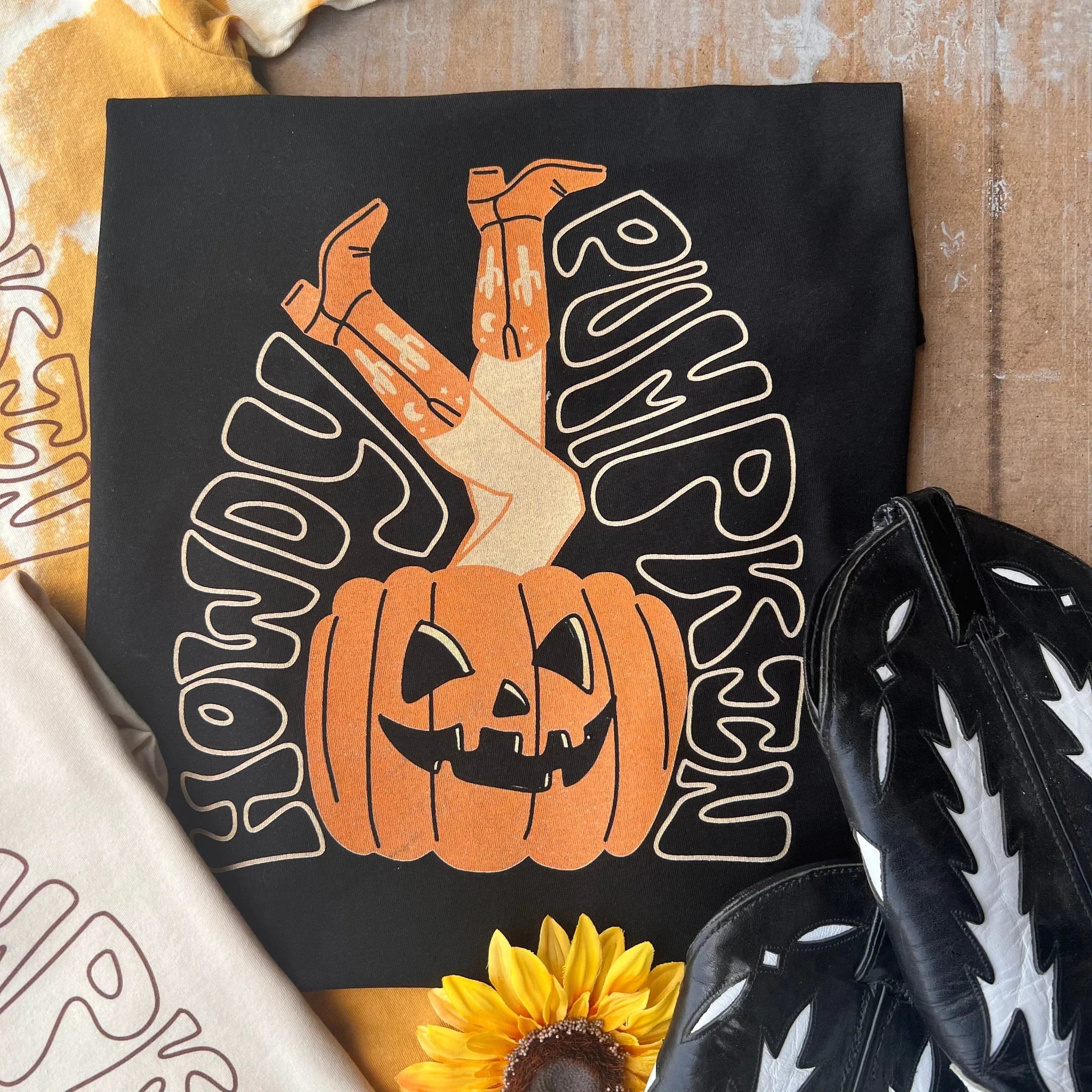 This is a black tee with white bubble letters that say howdy pumpkin and surround an orange pumpkin with cowgirl legs sticking out. It is pictured with black and white cowgirl boots and two other tees that are mostly cropped out of the photo.