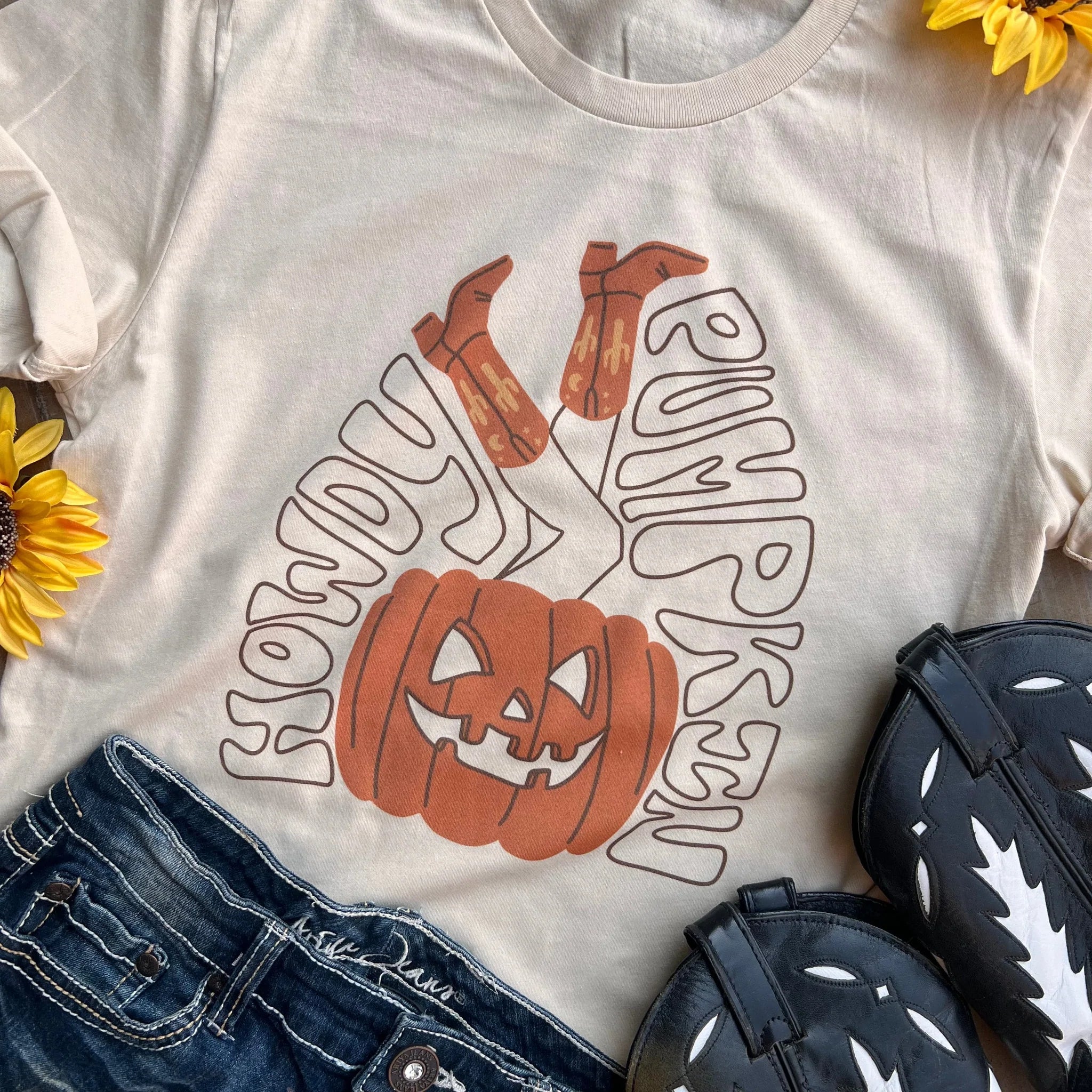 This is a cream tee that says howdy pumpkin in black bubble letters with a pumpkin with cowgirl legs sticking out of it. The words howdy pumpkin are surrounding the pumpkin and cowgirl legs. It is pictured with a pair of black and white cowgirl boots and a pair of jeans.