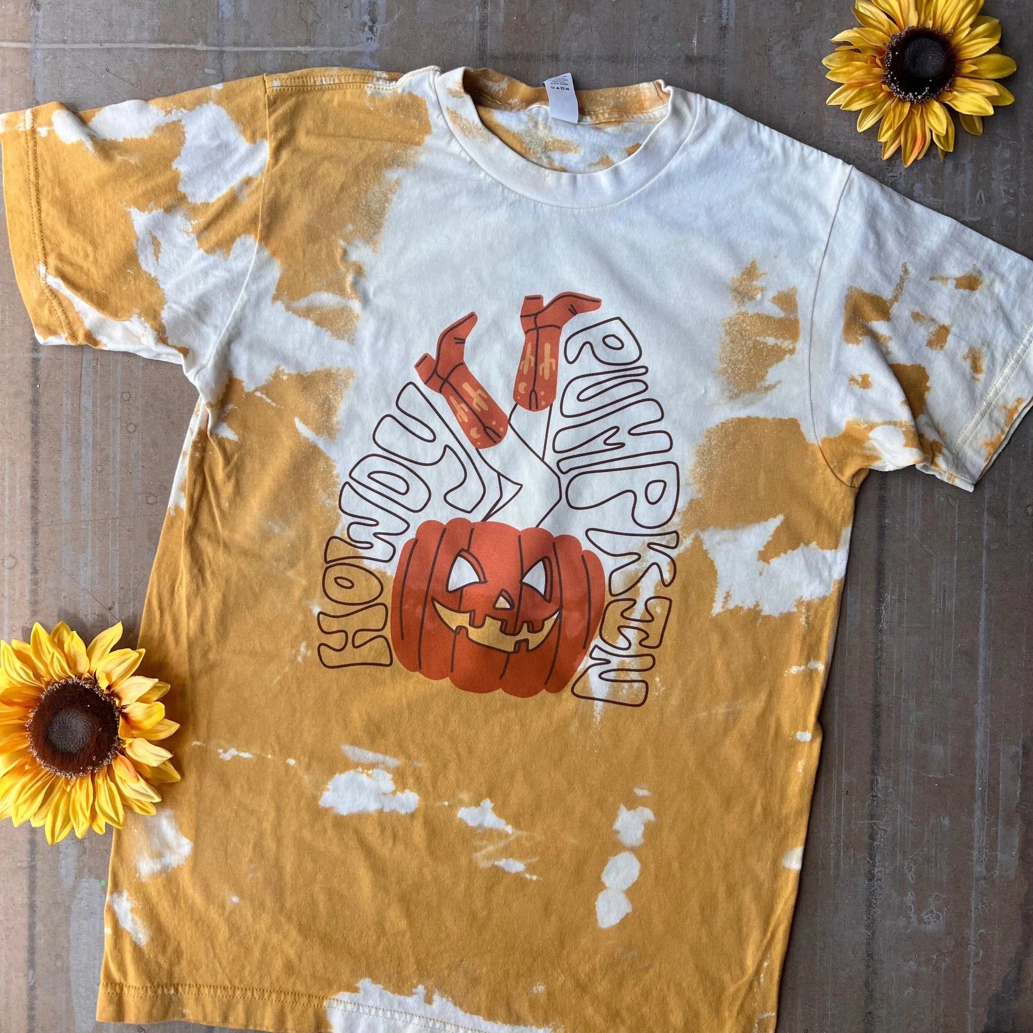 This is a mustard yellow tee that has been bleached. It says howdy pumpkin in black bubble letters with a pumpkin with cowgirl legs sticking out of it. The words howdy pumpkin are surrounding the pumpkin and cowgirl legs. There are also two sunflowers in the background of this picture.