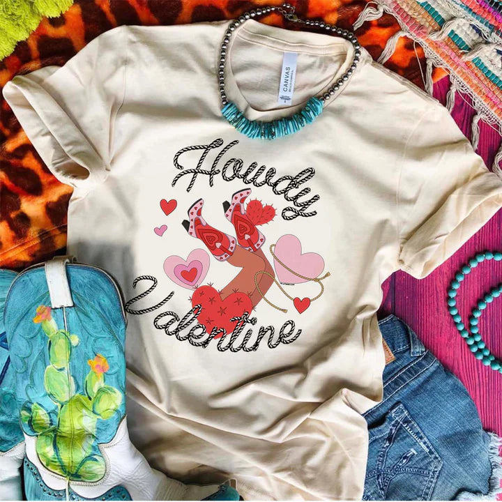 This crew neck cream tee is shown here as a flatlay with rolled sleeves on top of multicolored items along with denim shorts, a turquoise necklace, and turquoise boots with cactus. The graphic says "howdy valentine" in a cursive rope font. There are hearts, one being lassoed, and legs with cute pink and red boots up in the air centered between the "howdy valentine" font. 