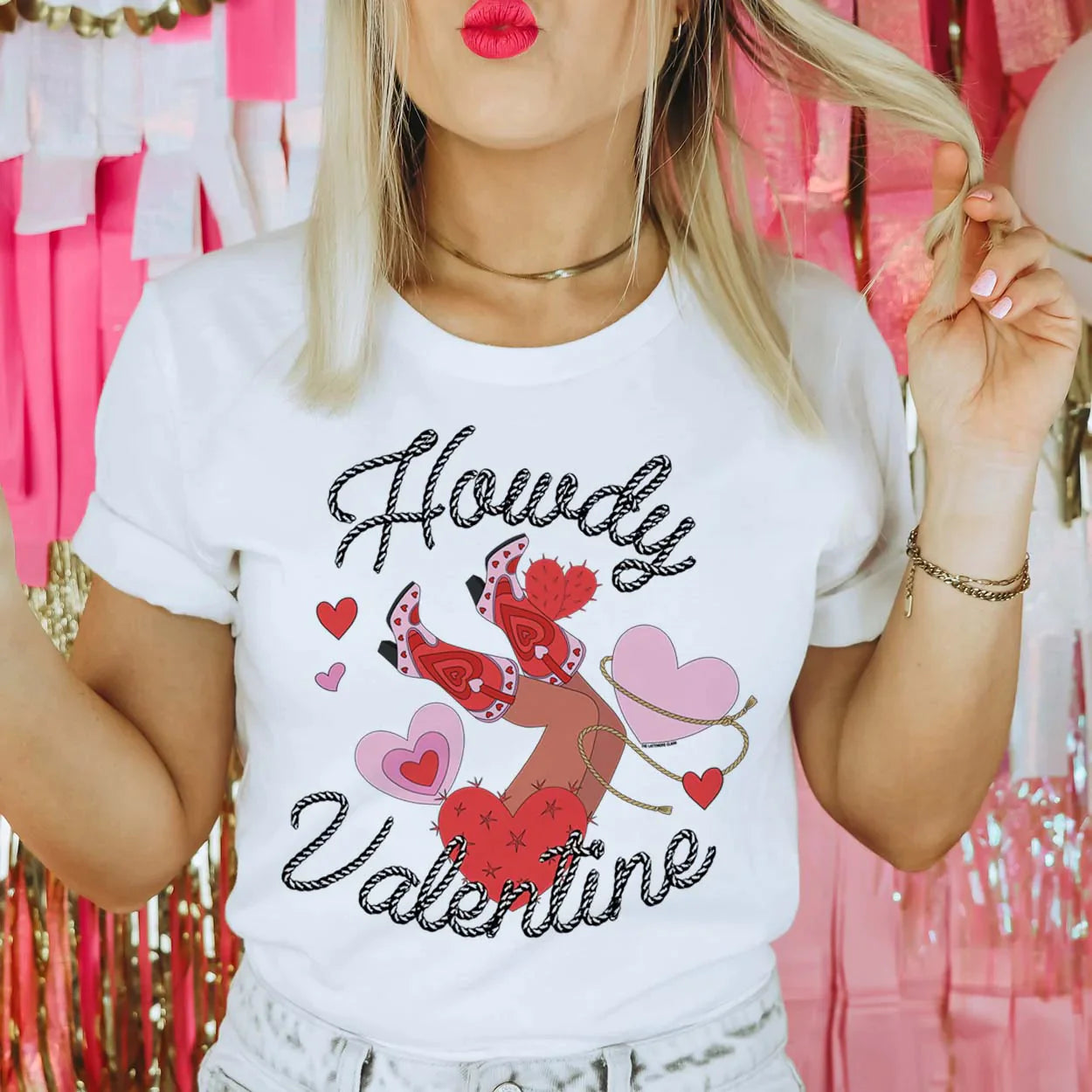 This crew neck white tee is shown here being modeled with rolled sleeves and gold jewelry and jeans.  The graphic says "howdy valentine" in a cursive rope font. There are hearts, one being lassoed, and legs with cute pink and red boots up in the air centered between the "howdy valentine" font. 