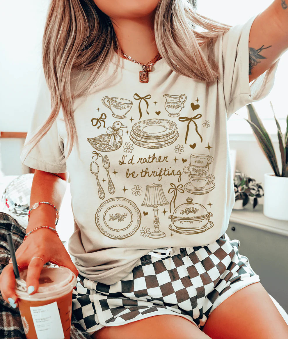 I’d Rather Be Thrifting graphic tee featuring vintage dishware, teacups, bows, and thrift inspired design on a cream shirt.
