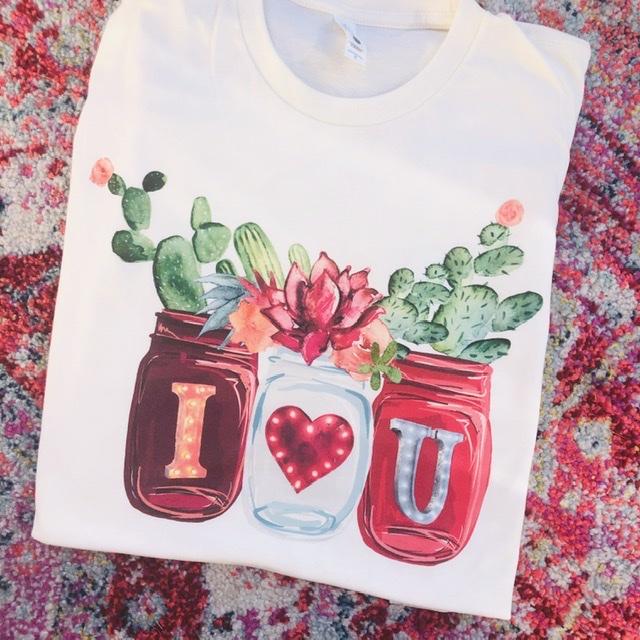 A white tee shirt folded to expose the mason jar graphic that have the letters "I (heart symbol) U." The jars have succulents growing out of the top. Tee shirt is pictured on top of a pink paisley rug.