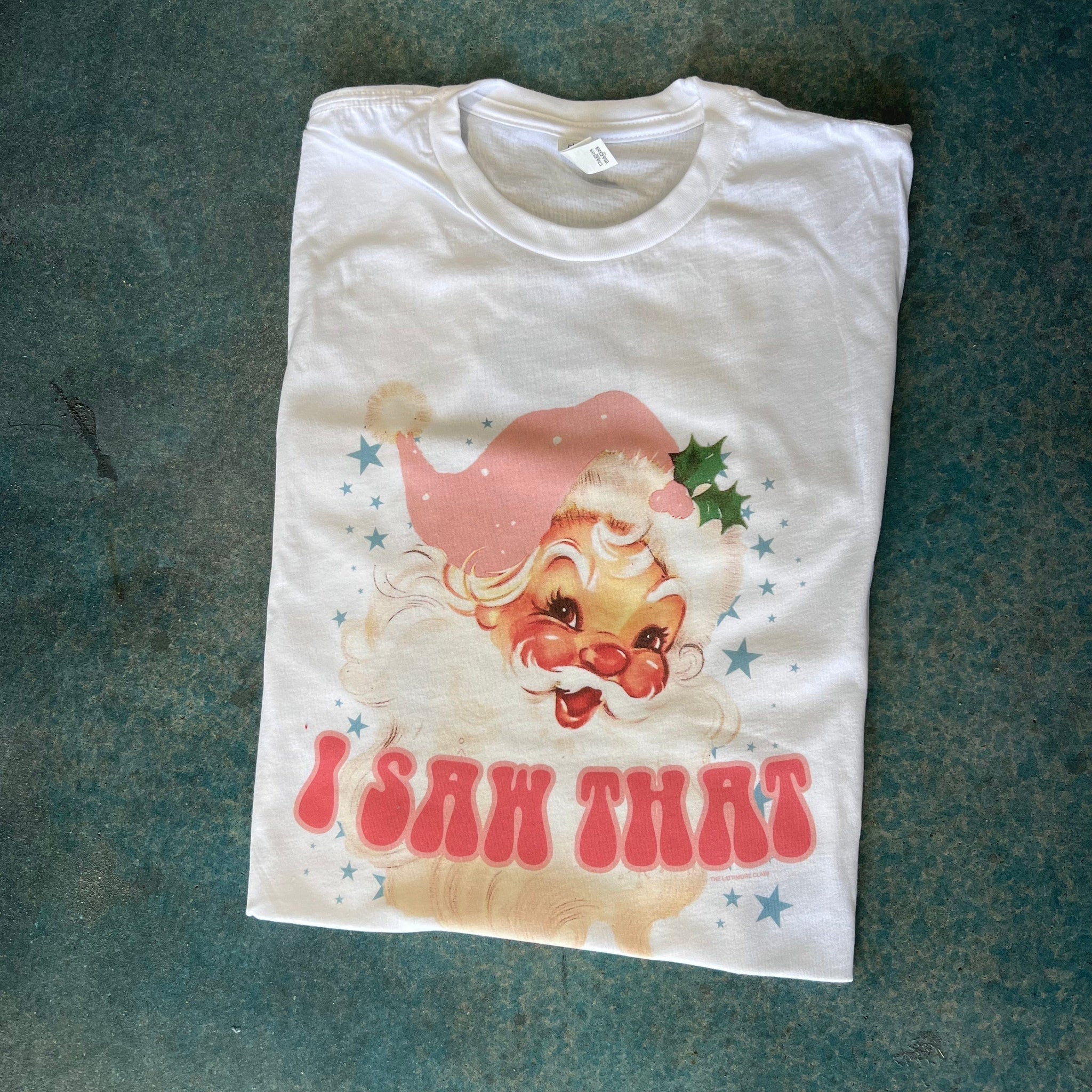 This white tee features a crew neckline, short sleeves, and a hand drawn graphic of a vintage Santa Clause with rosy cheeks wearing a light pink hat. There are blue stars around him of various sizes, along with the words "I SAW THAT" in a fun hot pink bubble font with light pink outline. It is shown here as a folded flatlay. 