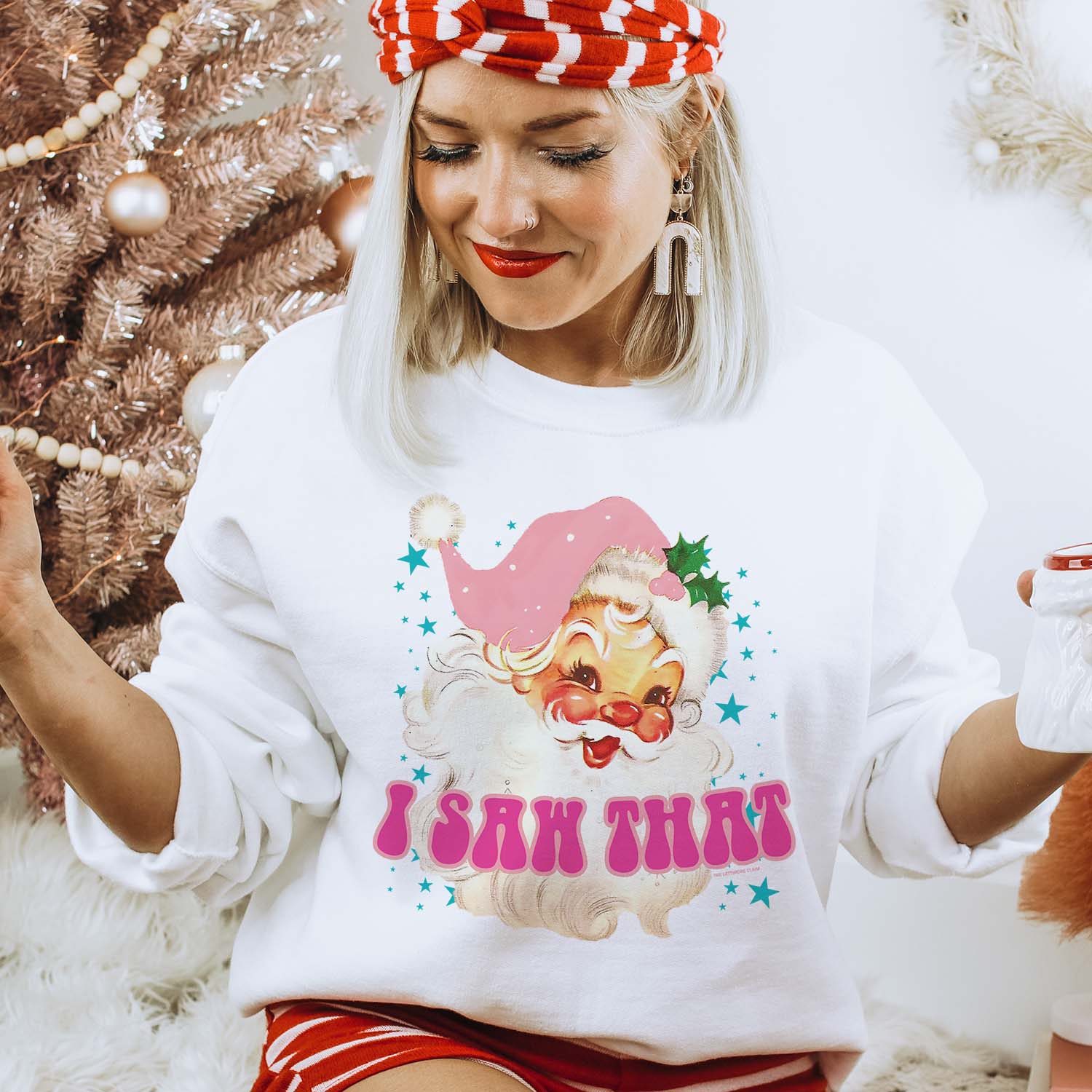 This white sweatshirt includes a crew neckline, long sleeves, and a hand drawn graphic of a vintage Santa Clause with rosy cheeks wearing a light pink hat. There are blue stars around him of various sizes, along with the words "I SAW THAT" in a fun hot pink bubble font with light pink outline. This sweatshirt is shown here being modeled with a pair of red and white striped shorts and matching headband, along with a pair of white dangle earrings.  