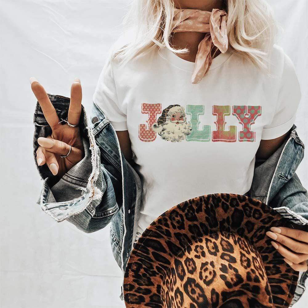 This white tee includes a crew neckline, short sleeves, and a graphic that says "Jolly" in a cute font. The model has this graphic tee styled with a denim jean jacket, neutral wild rag tied around the neck, and leopard print felt hat. 