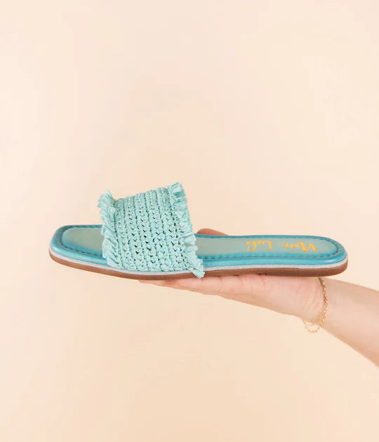 Turquoise woven sandal held in a hand against a beige background