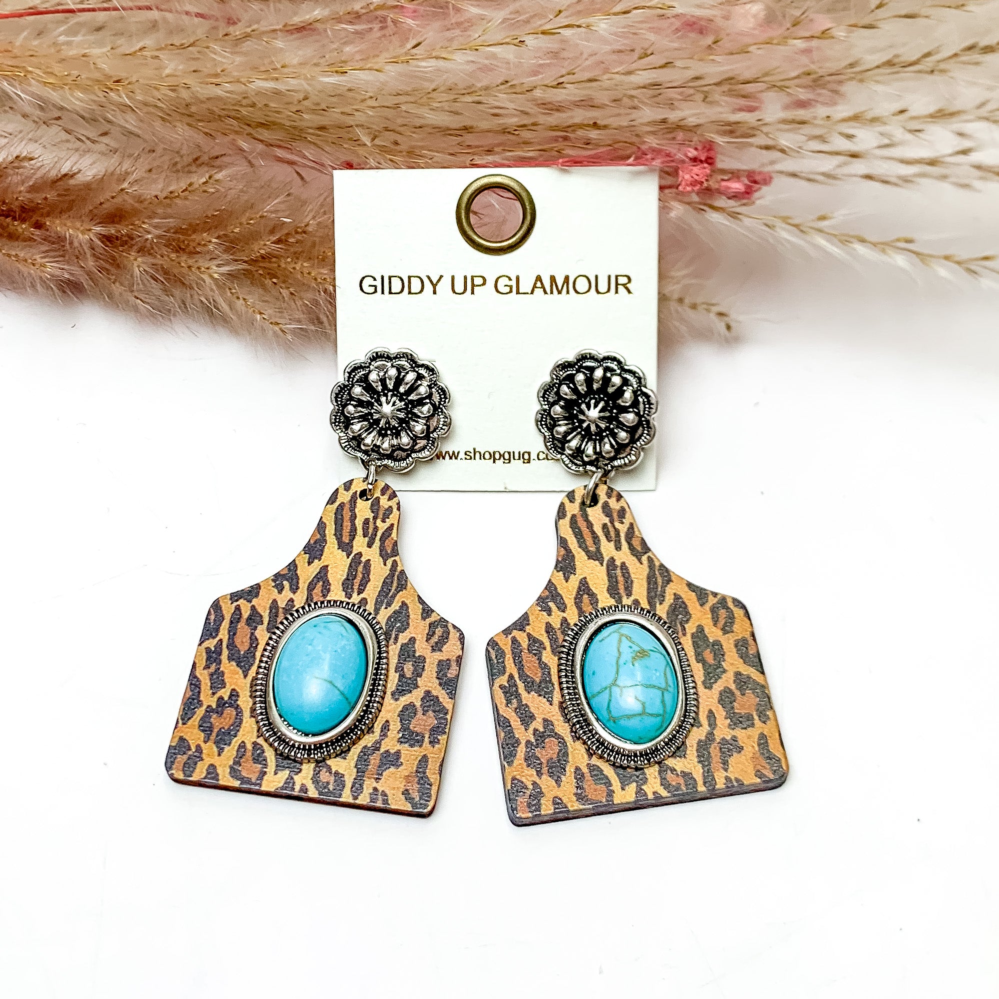 Leopard Cattle Tag Earrings With Turquoise Center Stone. These earrings are pictured on a white background with flowers behind for decoration.