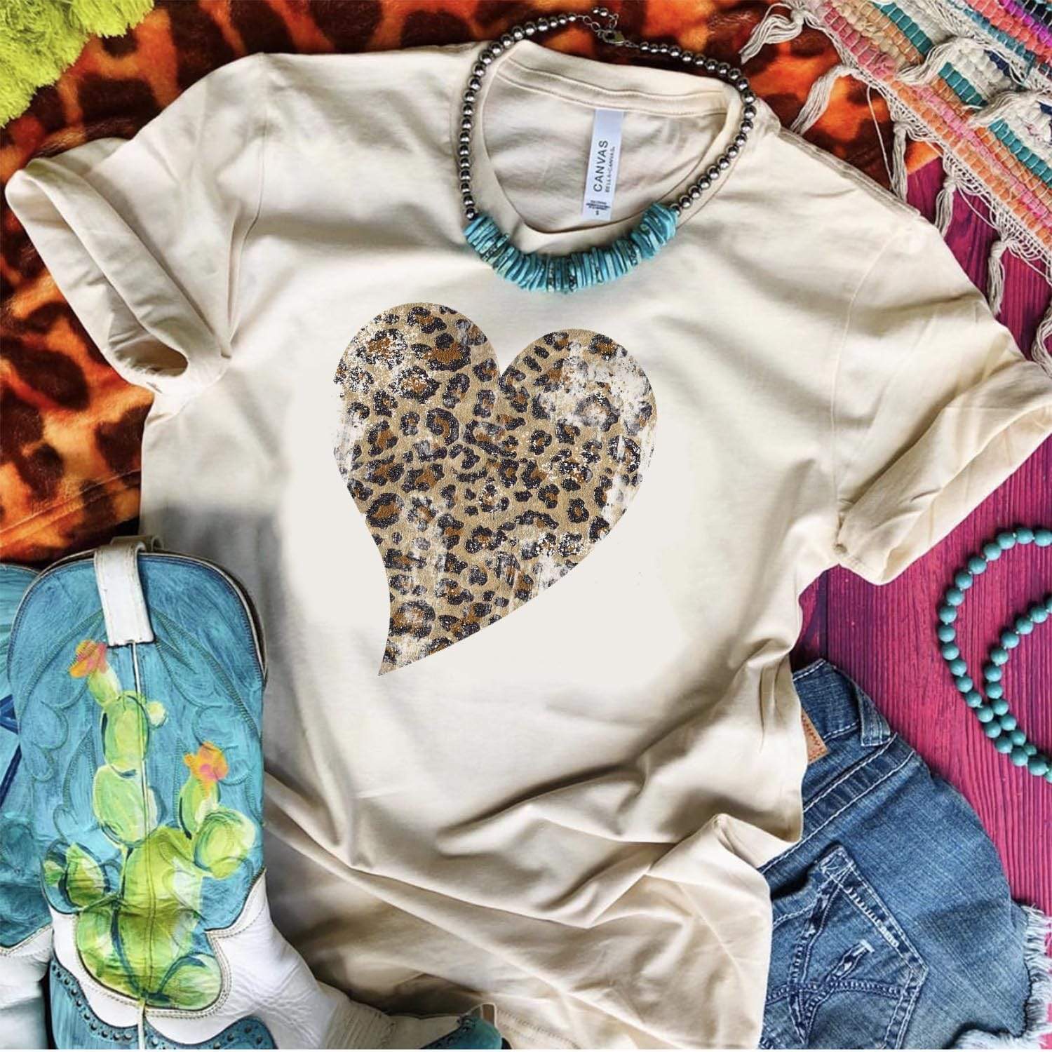 A cream color tee shirt laid on top of mix print rugs with cowgirl boots, a turquoise necklace, and denim shorts. The tee has a washed leopard print heart graphic.