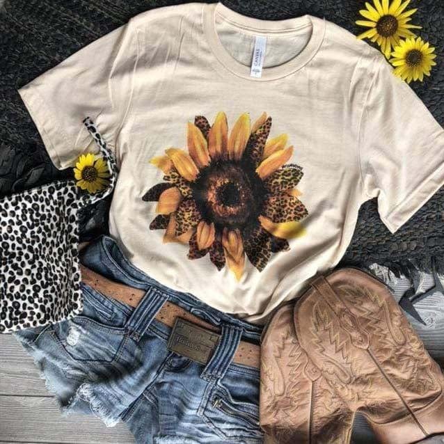 This is a cream t-shirt with a sunflower with some accent of leopard petals. It is pictured with a pair of blue jean shorts with a belt, brown cowgirl boots and a black and white leopard wristlet. The  outfit is placed on a black rug with 3 sunflowers surrounding the outfit. 