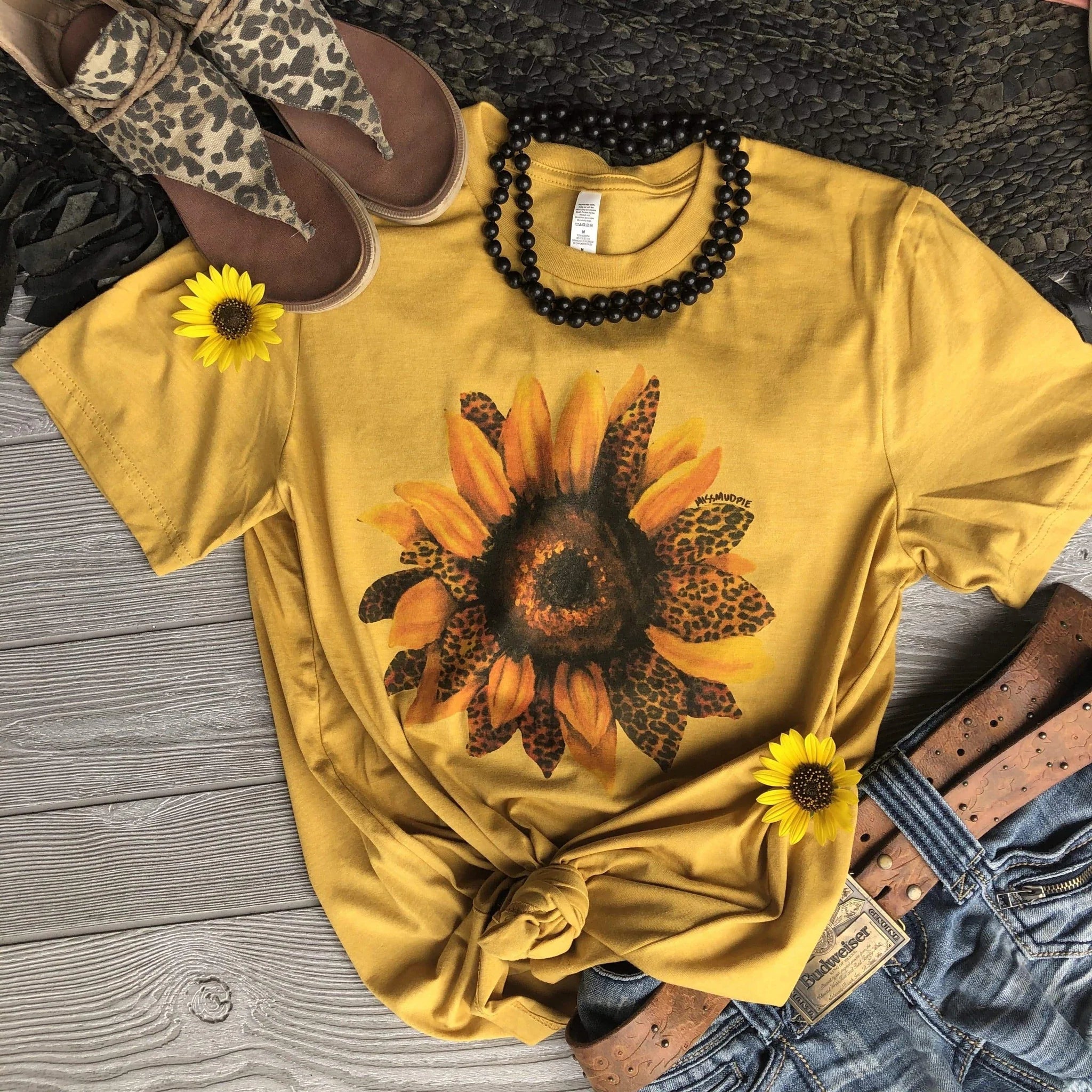 This is a mustard yellow t-shirt with a sunflower that has some leopard petals. This shirt is pictured with a pair of jeans and a brown belt, a black beaded necklace and some brown leopard sandals. 