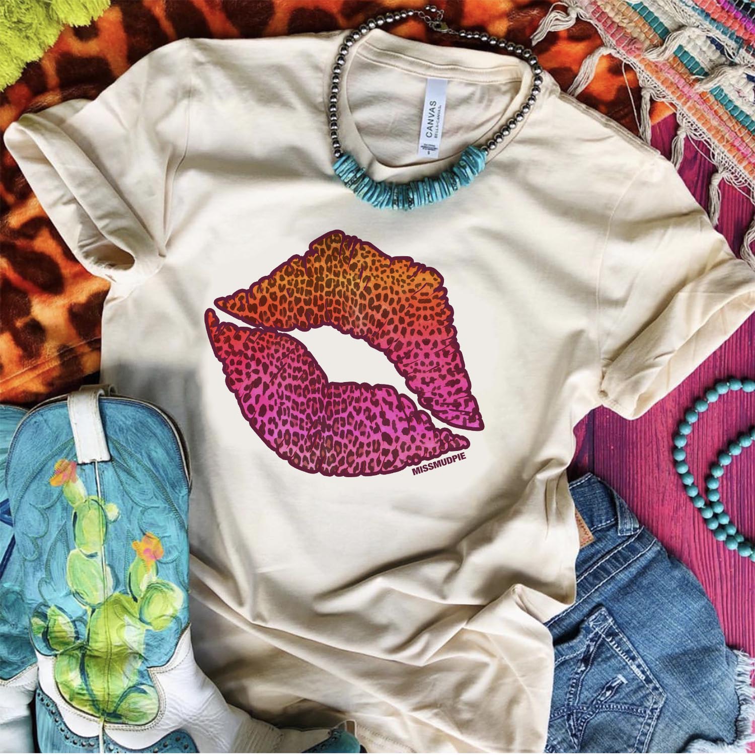 An ivory tee shirt laid on a purple wooden background with cowgirl boots, denim shorts, turquoise and silver necklace, and mix print rugs. The tee shirt has a graphic that is a leopard print kiss outline.