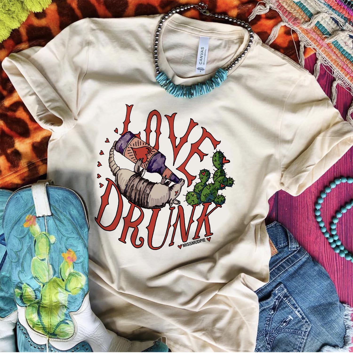 An ivory tee shirt laid on a purple wooden background with cowgirl boots, denim shorts, turquoise and silver necklace, and mix print rugs. The tee shirt has a graphic that says  "Love Drunk" with and armadillo drinking from a bottle.