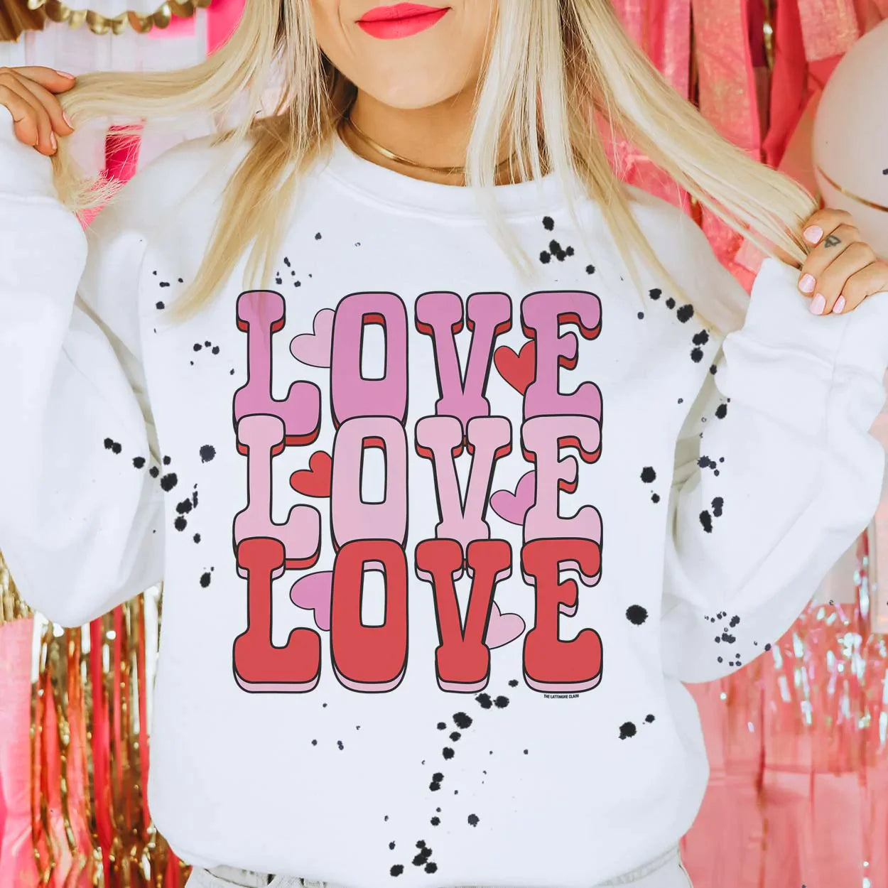 This white crew neck sweatshirt with black paint splatter is shown being modeled with jeans and a gold necklace. The graphic says "love love love" in hot pink, light pink, and red colored bubble font stacked. There are also hot pink, light pink, and red hearts throughout the graphic. 