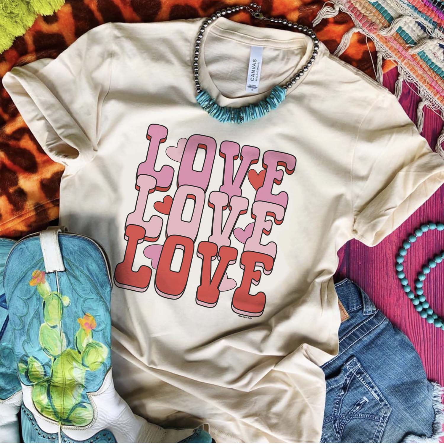 This cream tee is shown as a flatlay on a multi-colored background along with turquoise + cactus cowboy boots, denim shorts, and a turquoise and silver necklace. The graphic has the words "LOVE LOVE LOVE" in an ombre color pattern of hot pink, light pink, and red in all capital bubble font. There are also hearts randomly in between letters. This tee is also shown here with rolled sleeves. 