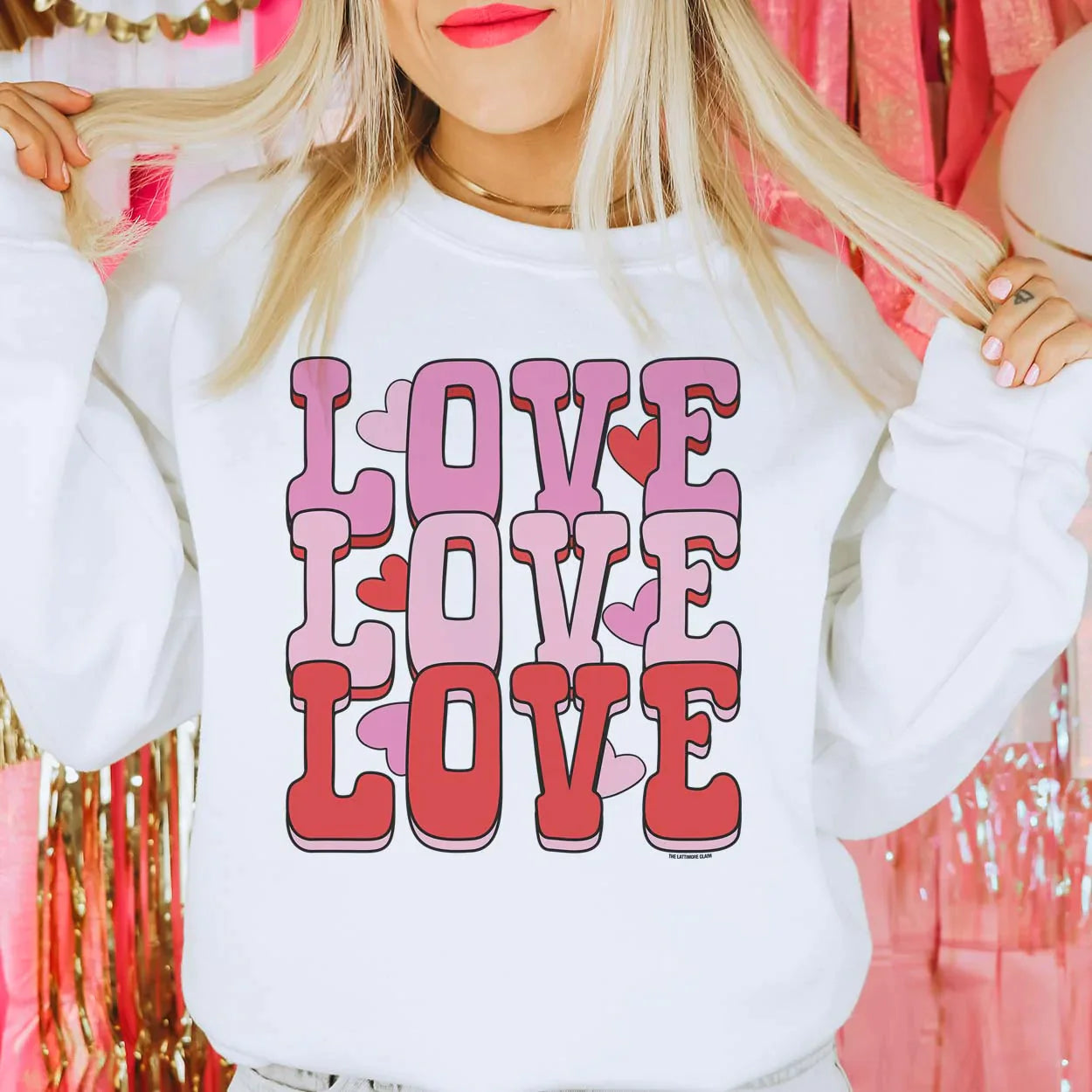 This white crew neck sweatshirt is shown being modeled with jeans and a gold necklace. The graphic says "love love love" in hot pink, light pink, and red colored bubble font stacked. There are also hot pink, light pink, and red hearts throughout the graphic. 