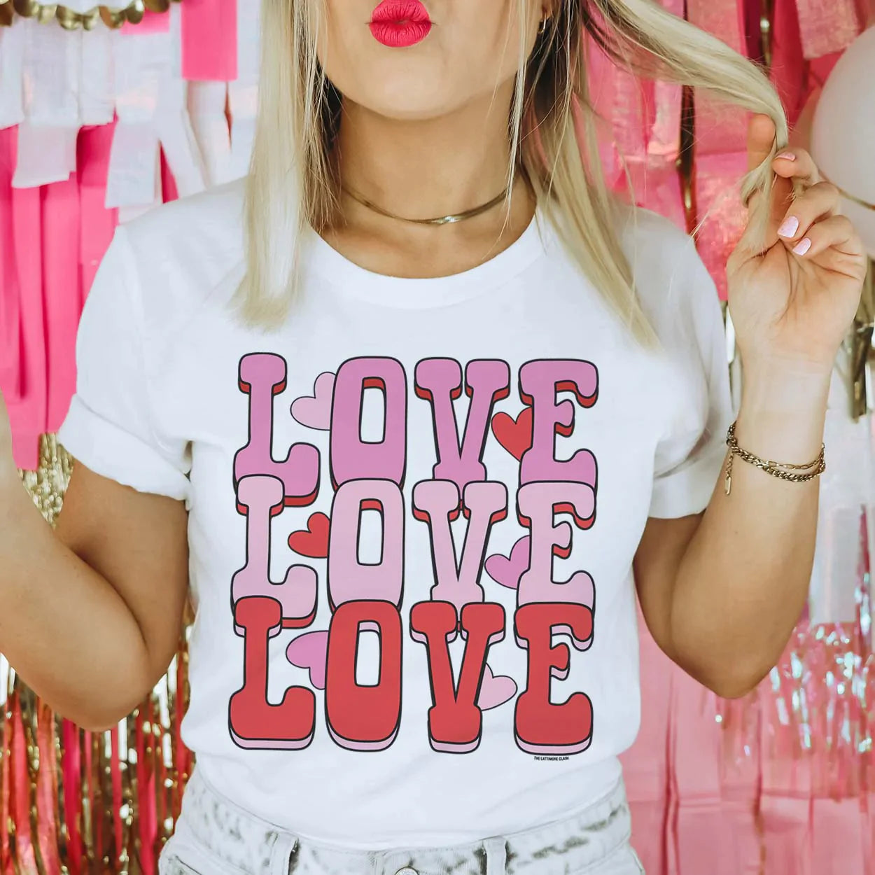 This crew neck, short sleeve white tee is being modeled with rolled sleeves and light wash denim jeans. The graphic says "love love love" in hot pink, light pink, and red colored bubble font, stacked on top of one another with hearts throughout. 