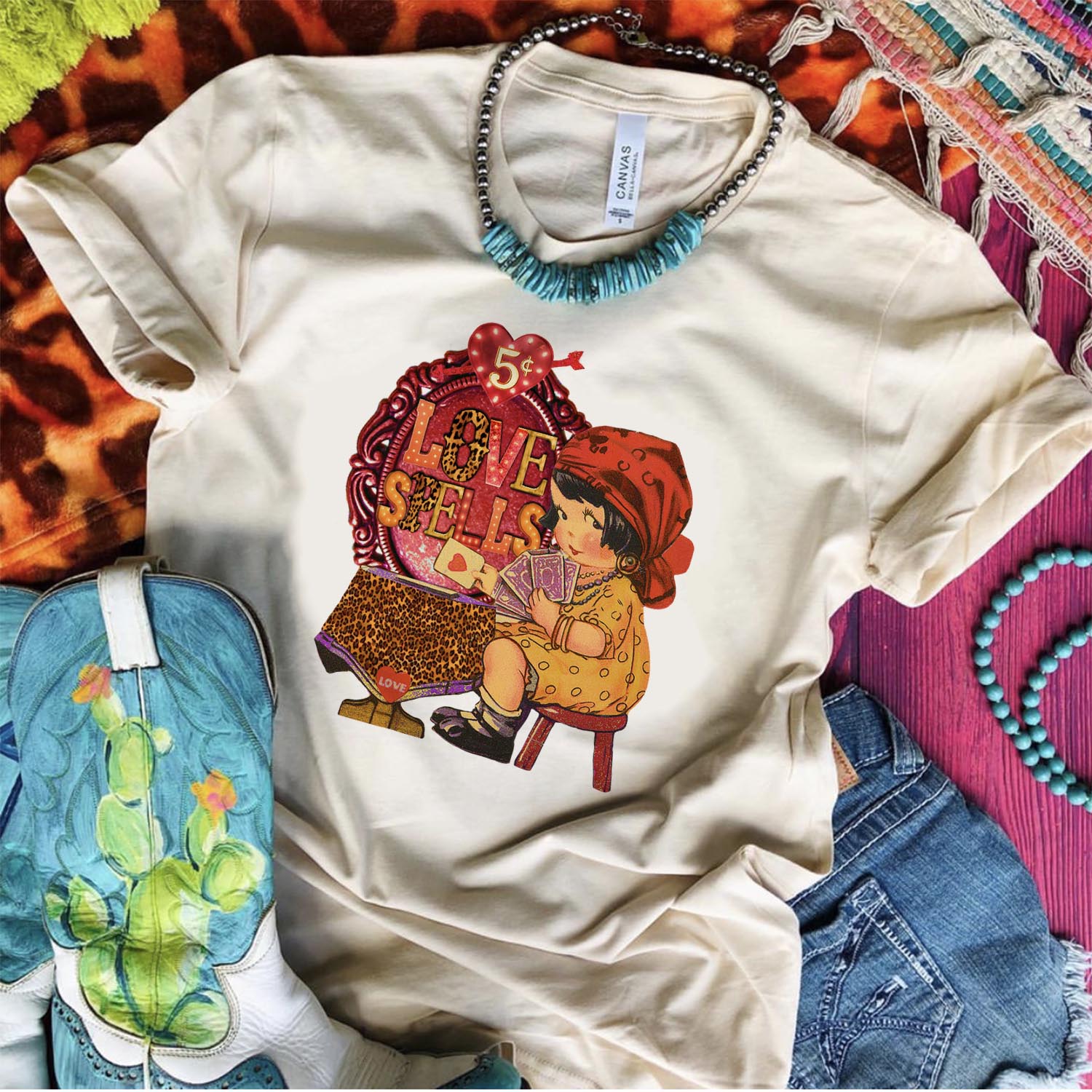 An ivory short sleeve teeshirt that has a gyspy graphic that says "Love Spells." Pictured on multicolored rugs with a turquoise necklace, cowgirl boots, and denim shorts.