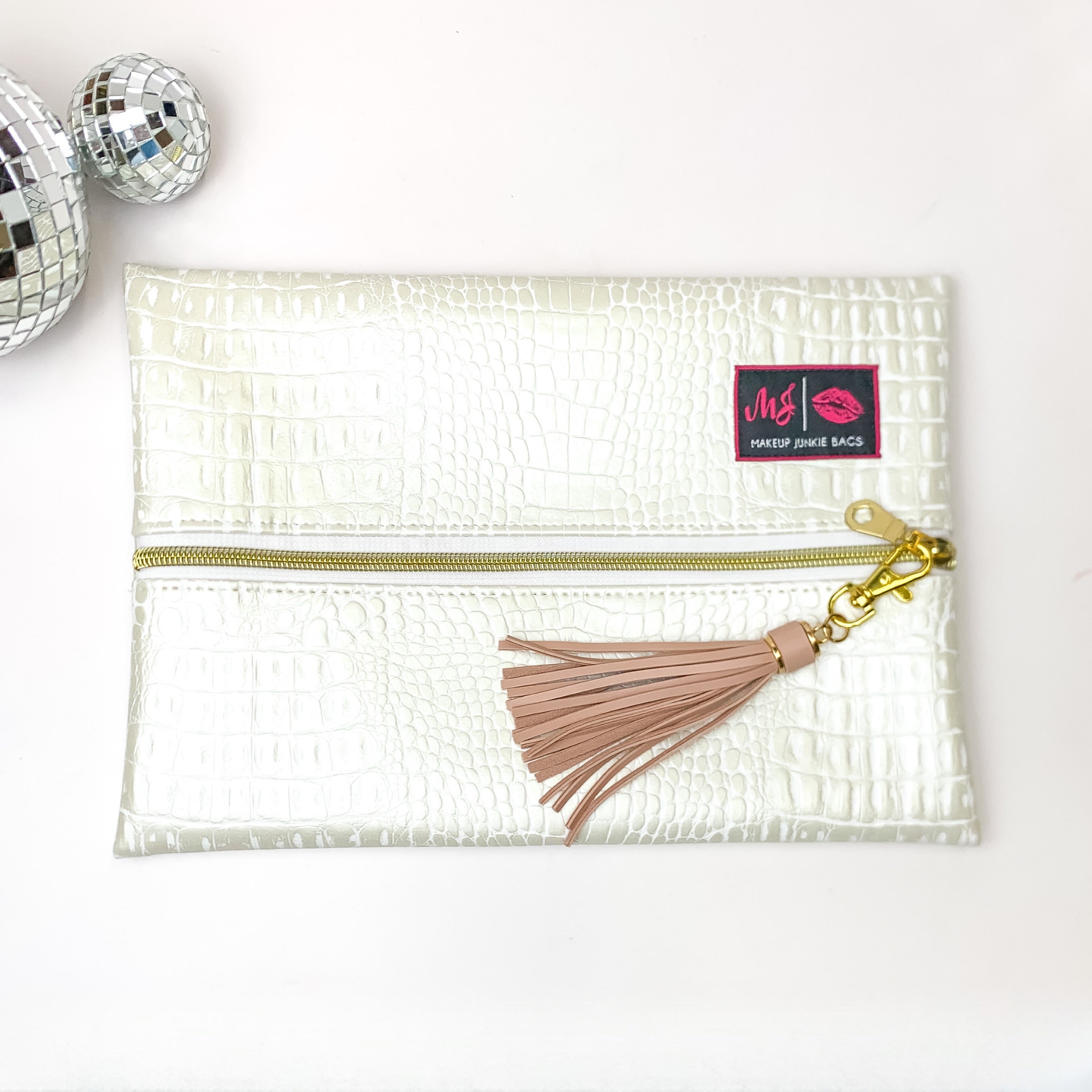 Pictured on a white background with disco balls at top is a lay flat bag in a pearl white gator print. This bag includes a middle zipper and a tassel.