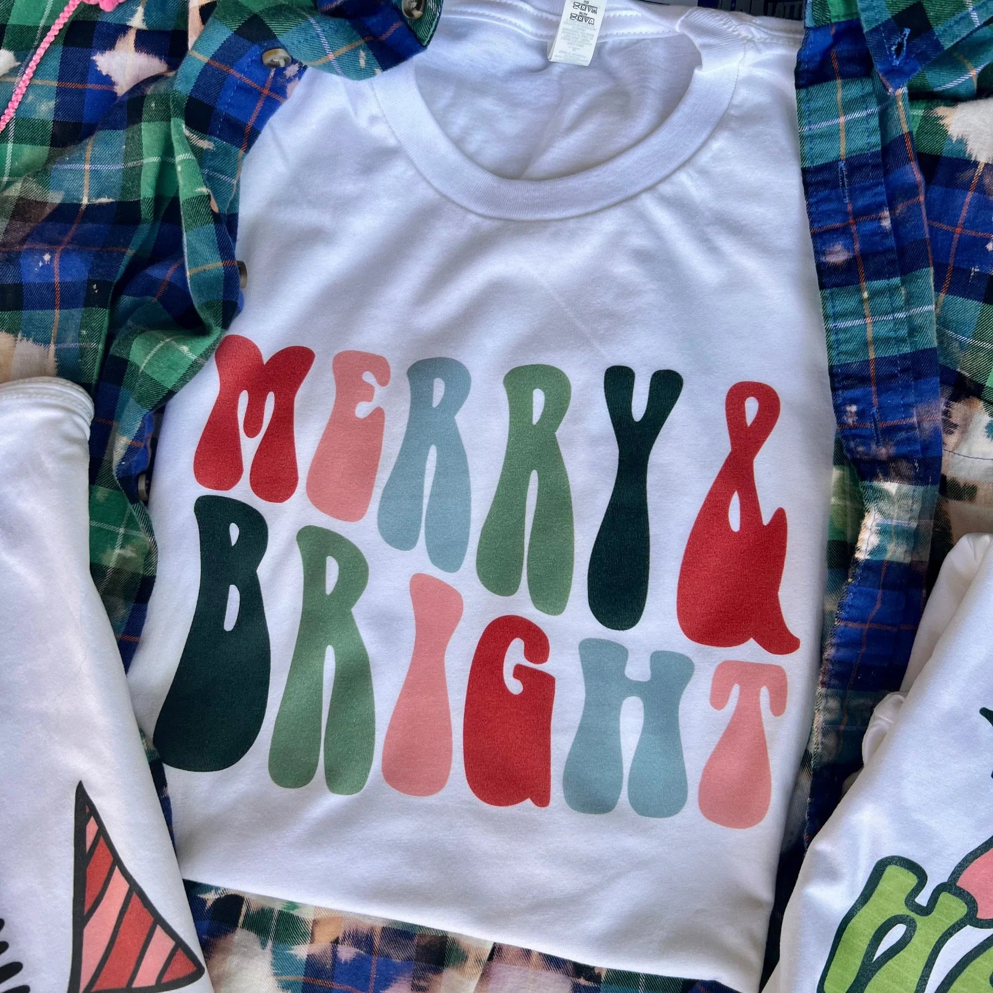 This white tee includes a crew neckline, short sleeves, and a cute Christmas saying "Merry & Bright" in a bubble font with different colors. This is shown here as a folded flat lay on top of a plaid button up shirt.