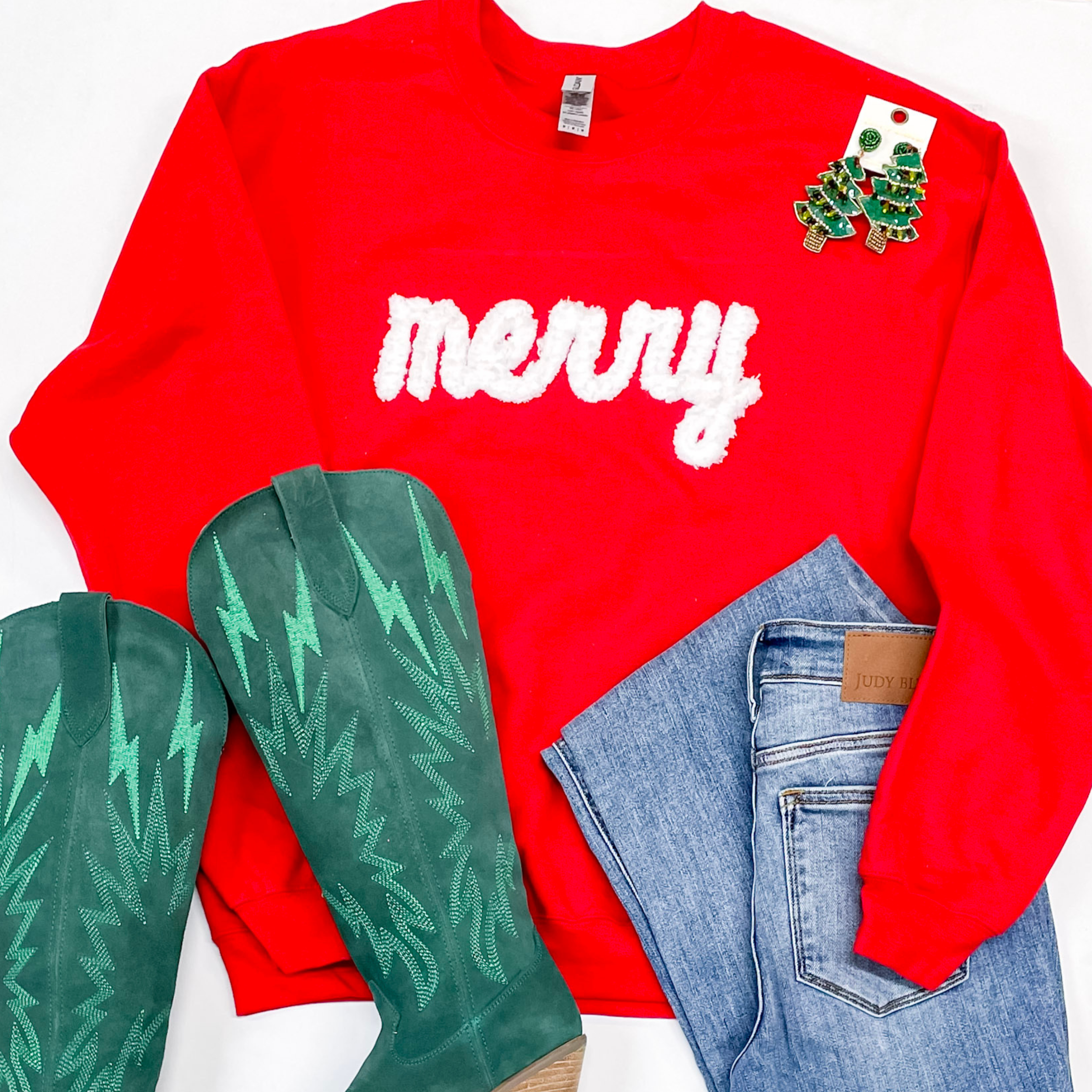 Photo features red sweatshirt with the word "Merry" across the front. This is paired with Judy Blue jeans, green Dingo boots, and green earrings all on a white background. 