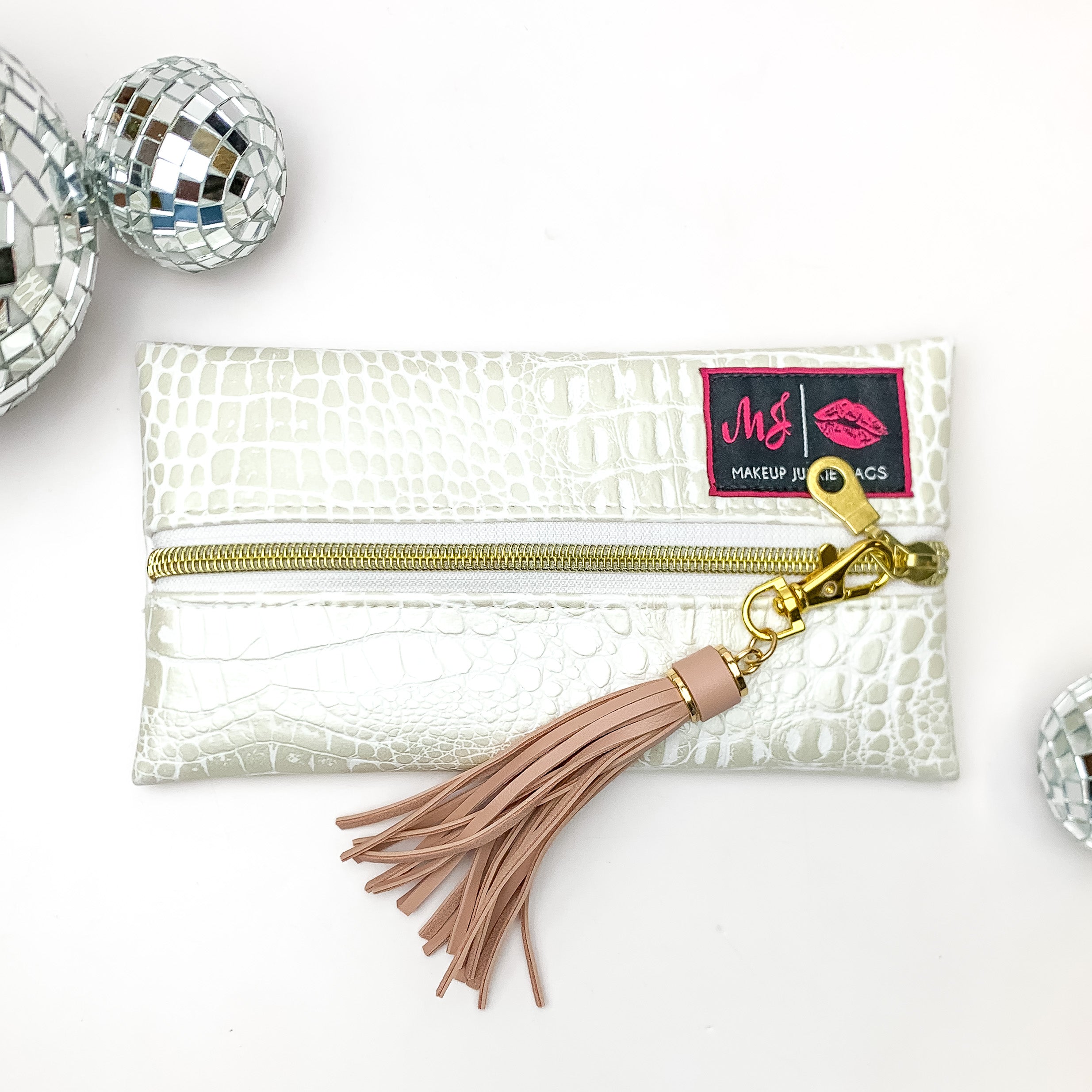 Pictured on a white background with disco balls at top is a mini lay flat bag in a pearl white gator print. This bag includes a middle zipper and a tassel. 