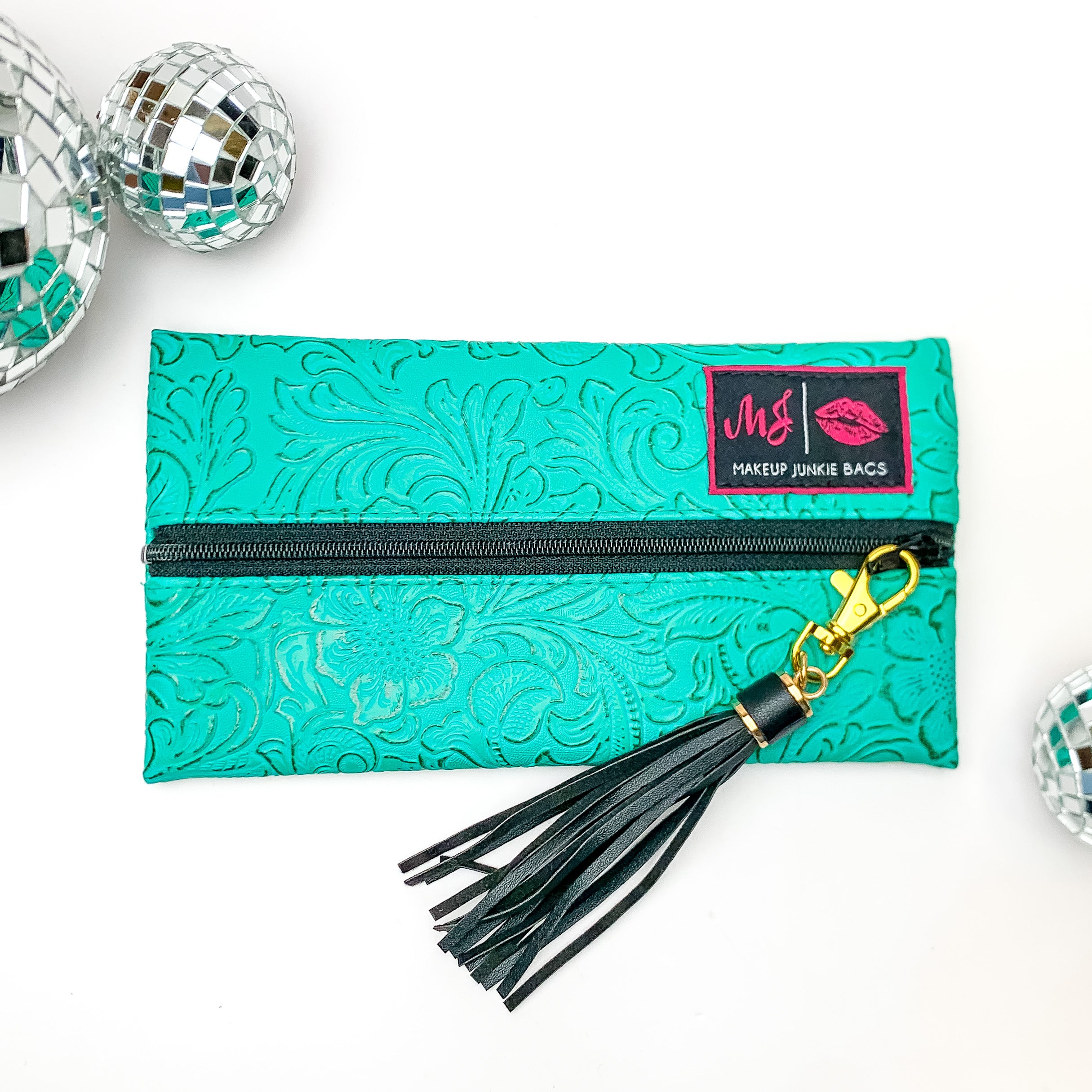 Pictured on a white background with disco balls at top is a mini lay flat bag in a turquoise leather tooled print. This bag includes a middle zipper and a tassel. 
