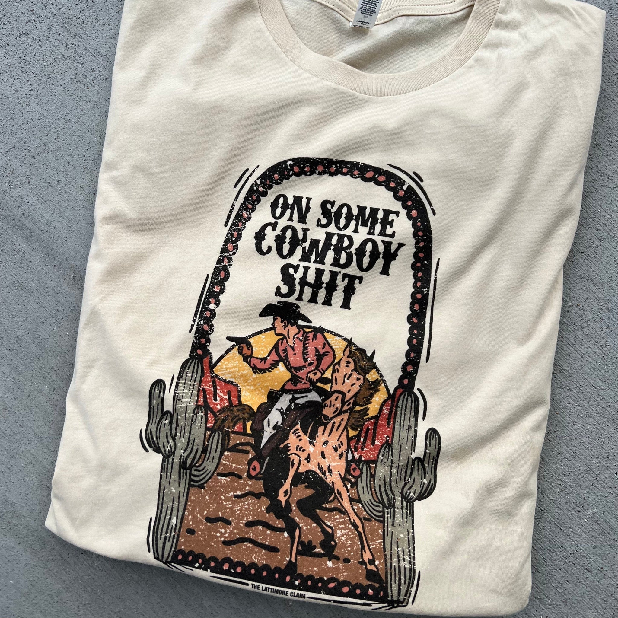 A white short sleeve crewneck tee with a graphic of a cowboy and his horse in the desert. The text "On some cowboy sh*t" is centered above the graphic. The item is pictured on a heather grey background