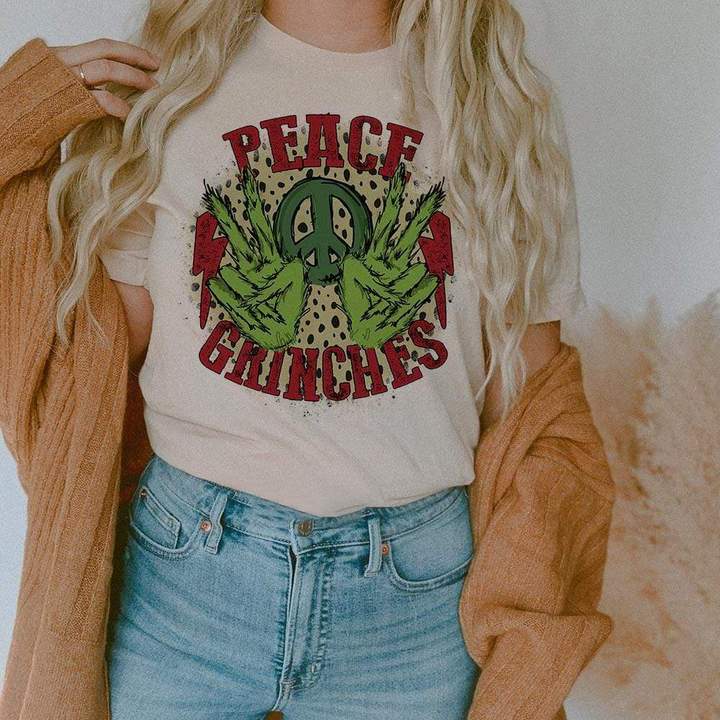 This cream tee includes a crew neckline, short sleeves, and a cute Christmas graphic that says "Peace Grinches" in red font, two Grinch hands holding up the peace sign, a green peace sign between the hands, and two red lightning bolts on the outside of the Grinch hands. This graphic also has a leopard print behind it. The model has this graphic tee styled with a camel cardigan and light wash denim jeans. 