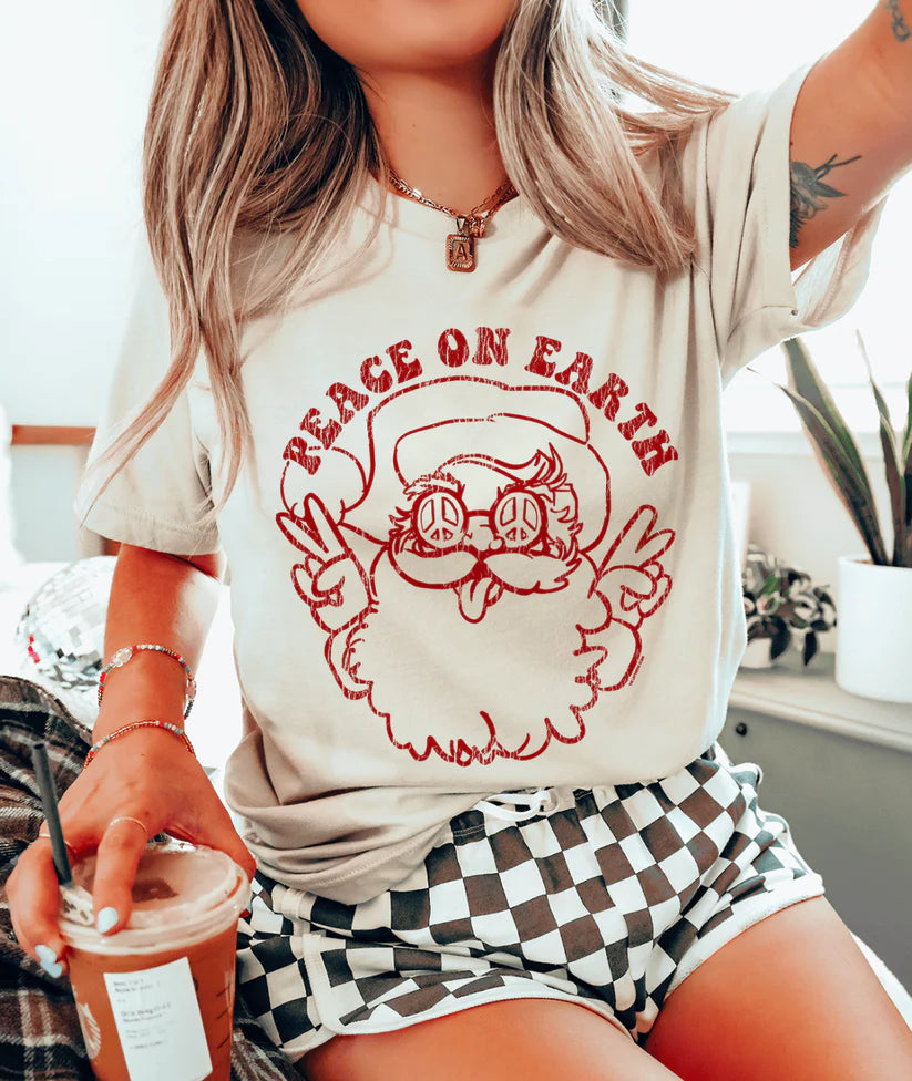 Cream tee with red “Peace on Earth” Santa graphic featuring peace sign sunglasses and retro lettering.
