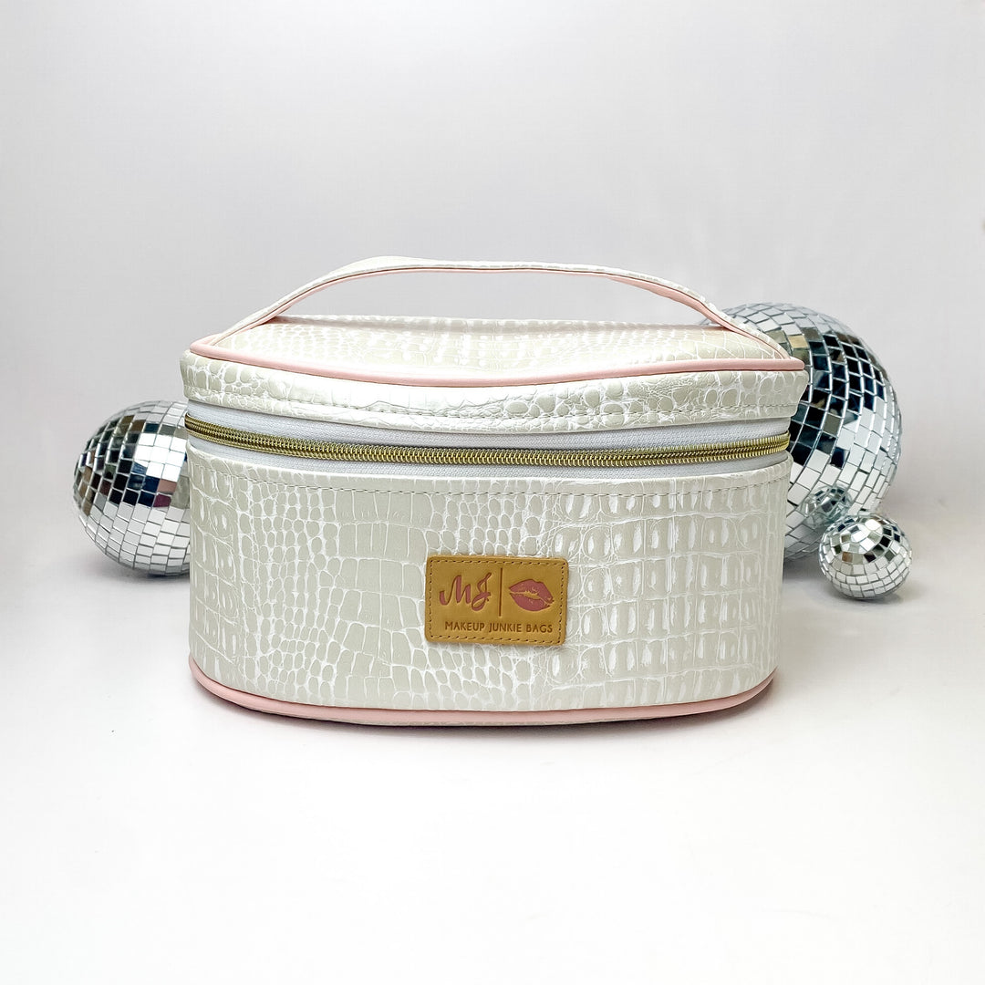 Pictured on a white background with disco balls in the background is a traincase with a top handle in a pearl white gator print. This bag includes a middle zipper and a tassel.