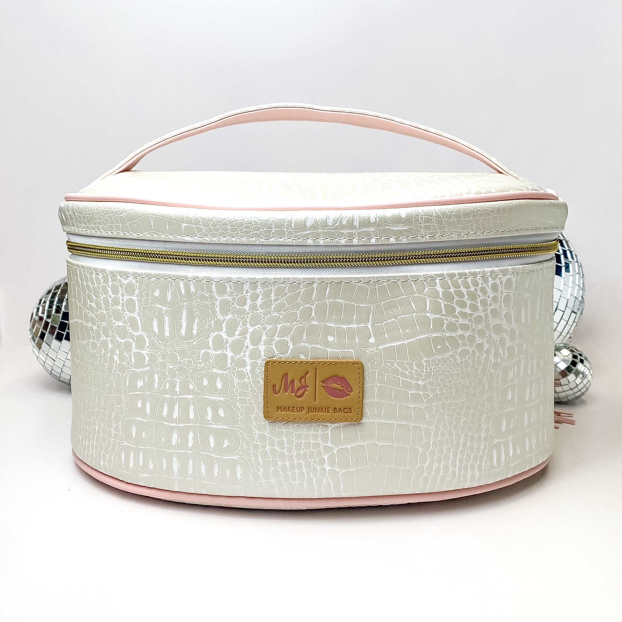 Pictured on a white background with disco balls in the background is a traincase with a top handle in a pearl white gator print. This bag includes a middle zipper and a tassel.