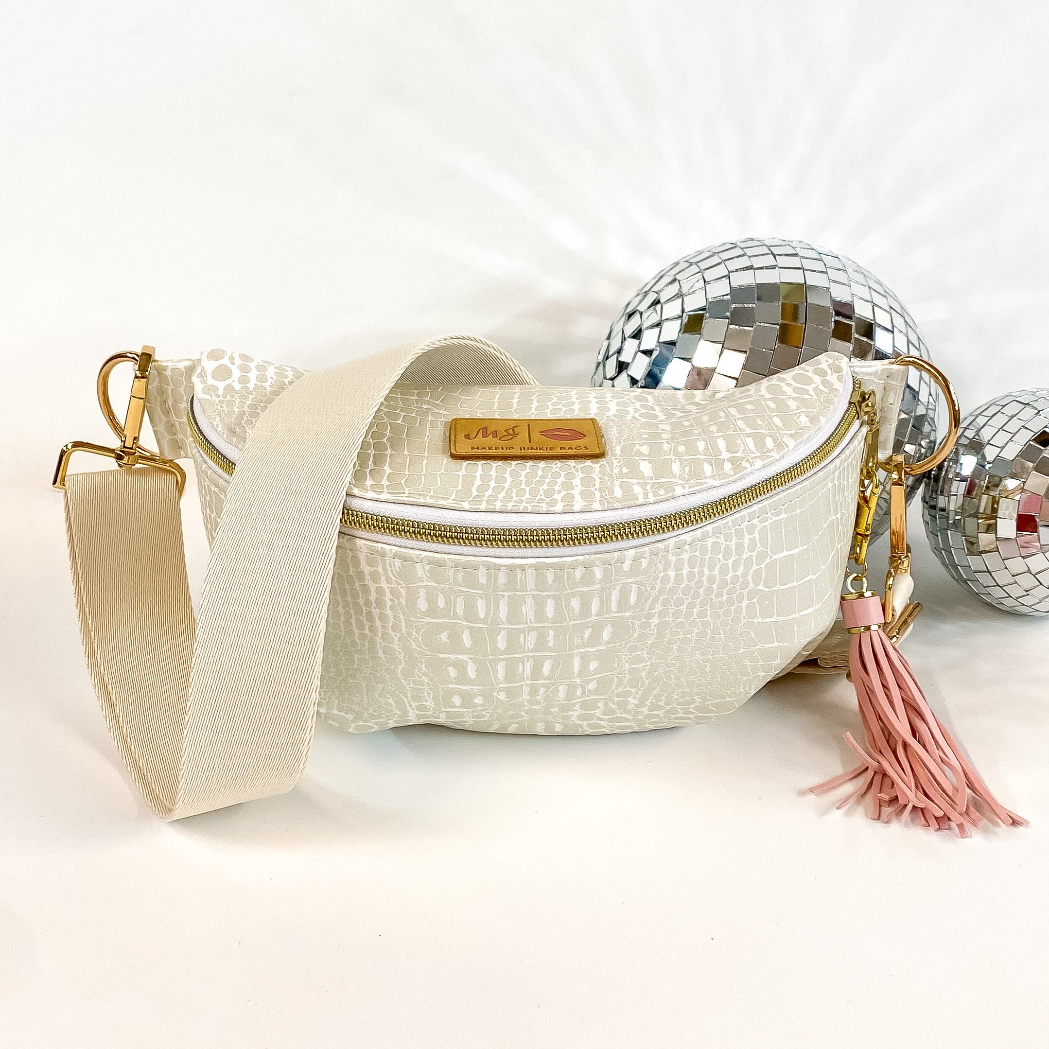 Pictured on a white background with disco balls in the background is a sidekick bag in a pearl white gator print. This bag includes a middle zipper and a tassel.