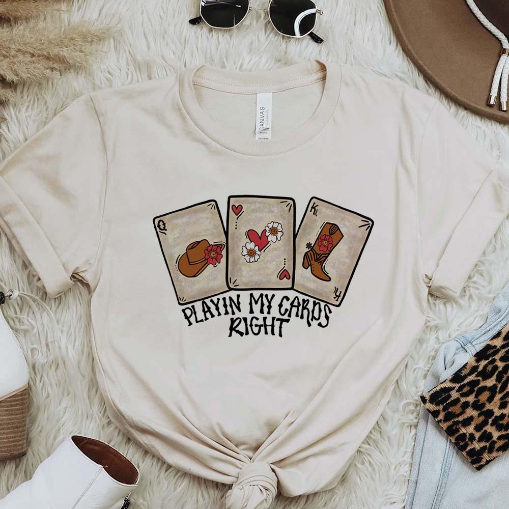 A cream colored tee shirt with playing cards across the front and says "Playin' My Cards Right." The tee shirt is pictured on a fur background with white booties, a brown hat, and sunglasses.
