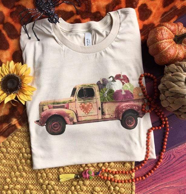 This is a cream graphic tee with a truck in the colors rusty cream, green and purple. The truck has pumpkins in the bed of the truck. The truck also has a lite up heart on the side of it. The background of the picture includes two decorative pumpkins, a sunflower and a glittered spider.