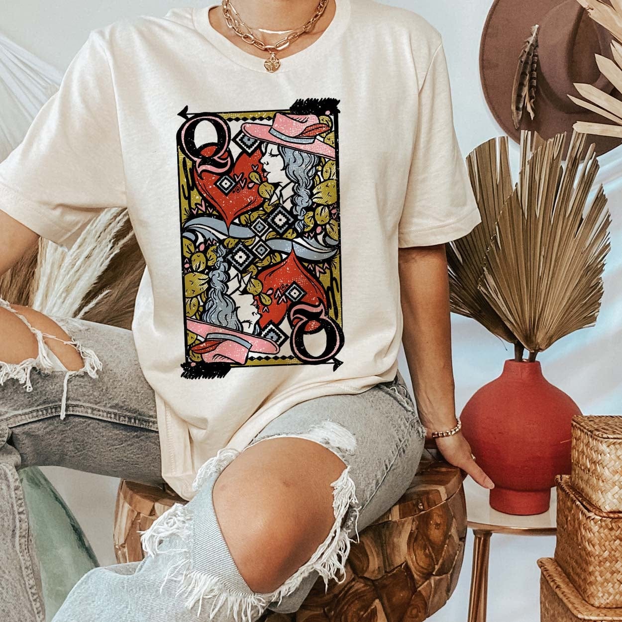Model is wearing a cream colored tee with a Queen of Hearts playing card printed on the front. Model has it paired with distressed jeans and gold jewelry.