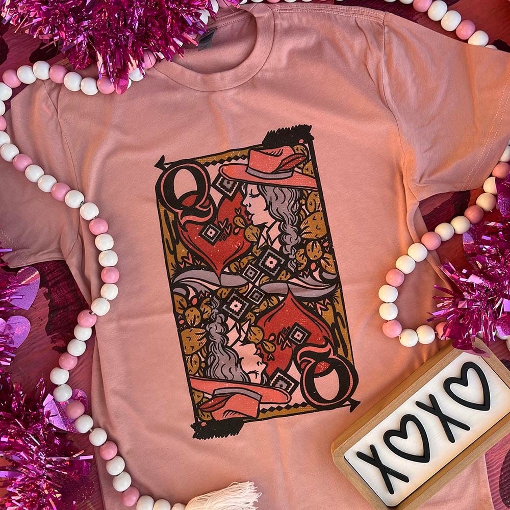 A desert rose tee shirt laid out on a wood background, pictured with pink and white beads and a "XOXO" wooden sign. This tee shirt has a graphic of a Queen of Hearts playing card on it.