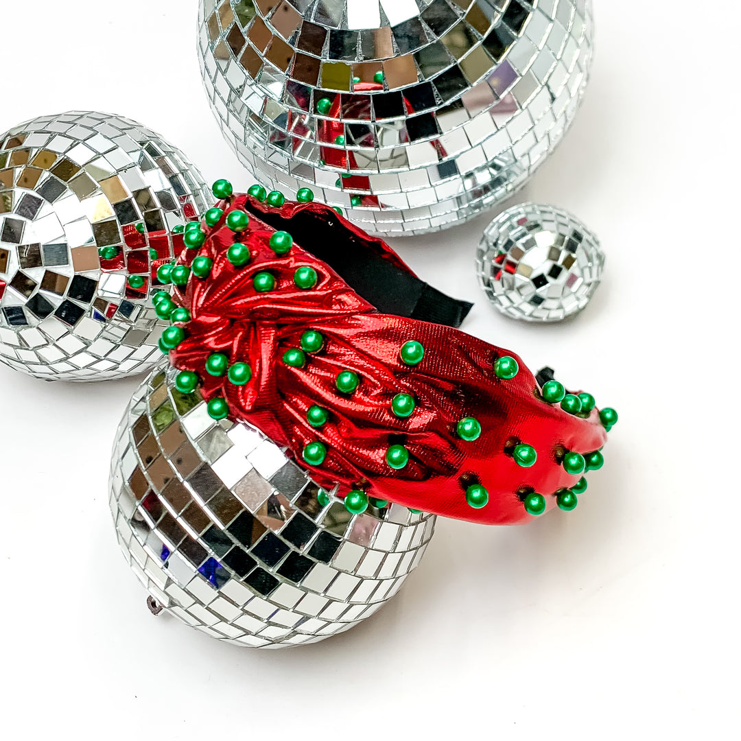 Top of the Charts Red Headband With Green Pearls. This headband is pictured on a white background and is surrounded by disco balls. 
